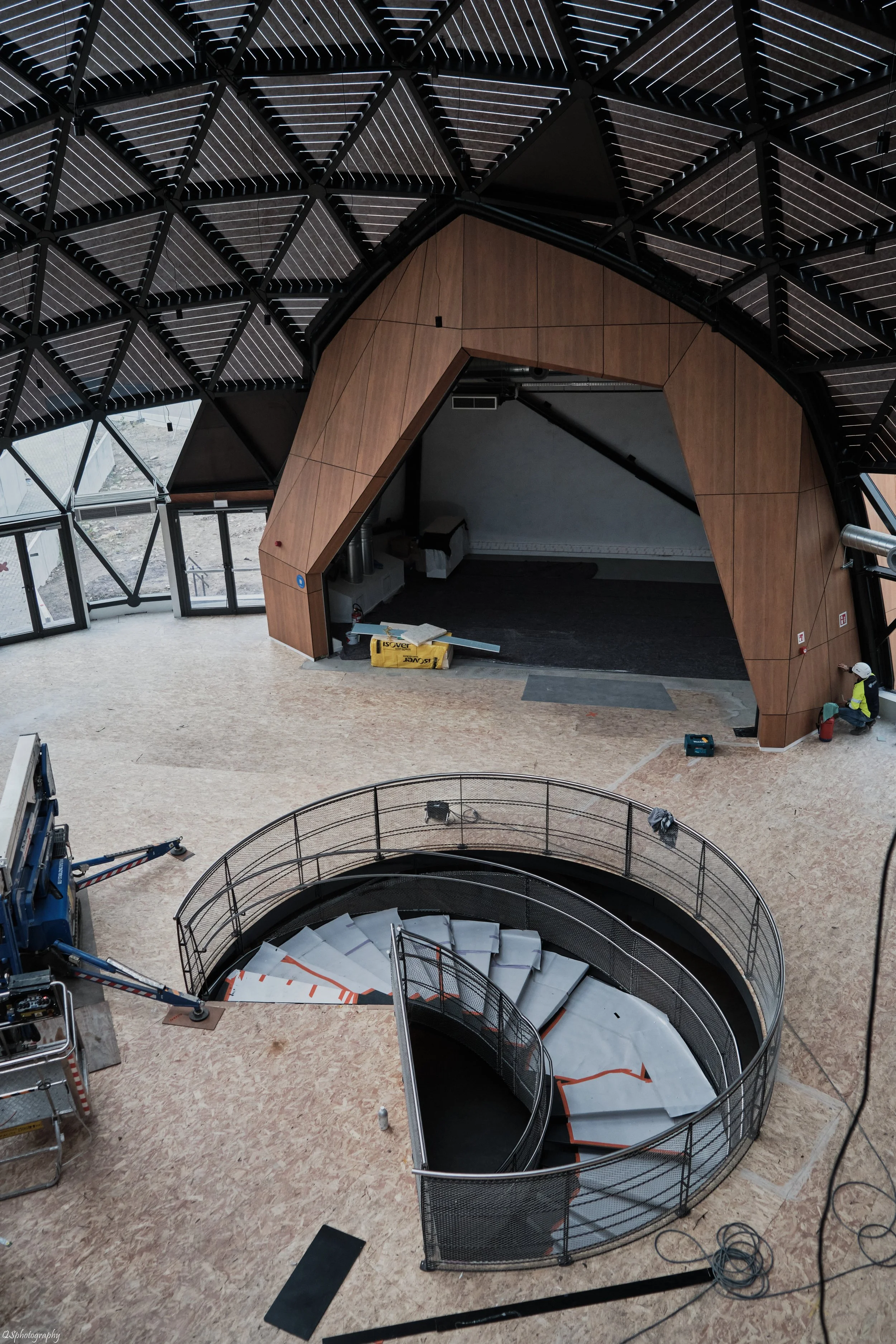 Vue intérieure d'un bâtiment en construction, avec un escalier en spirale non terminé au centre et une scène de chantier avec des travailleurs et des équipements. | QSphotography | Namur
