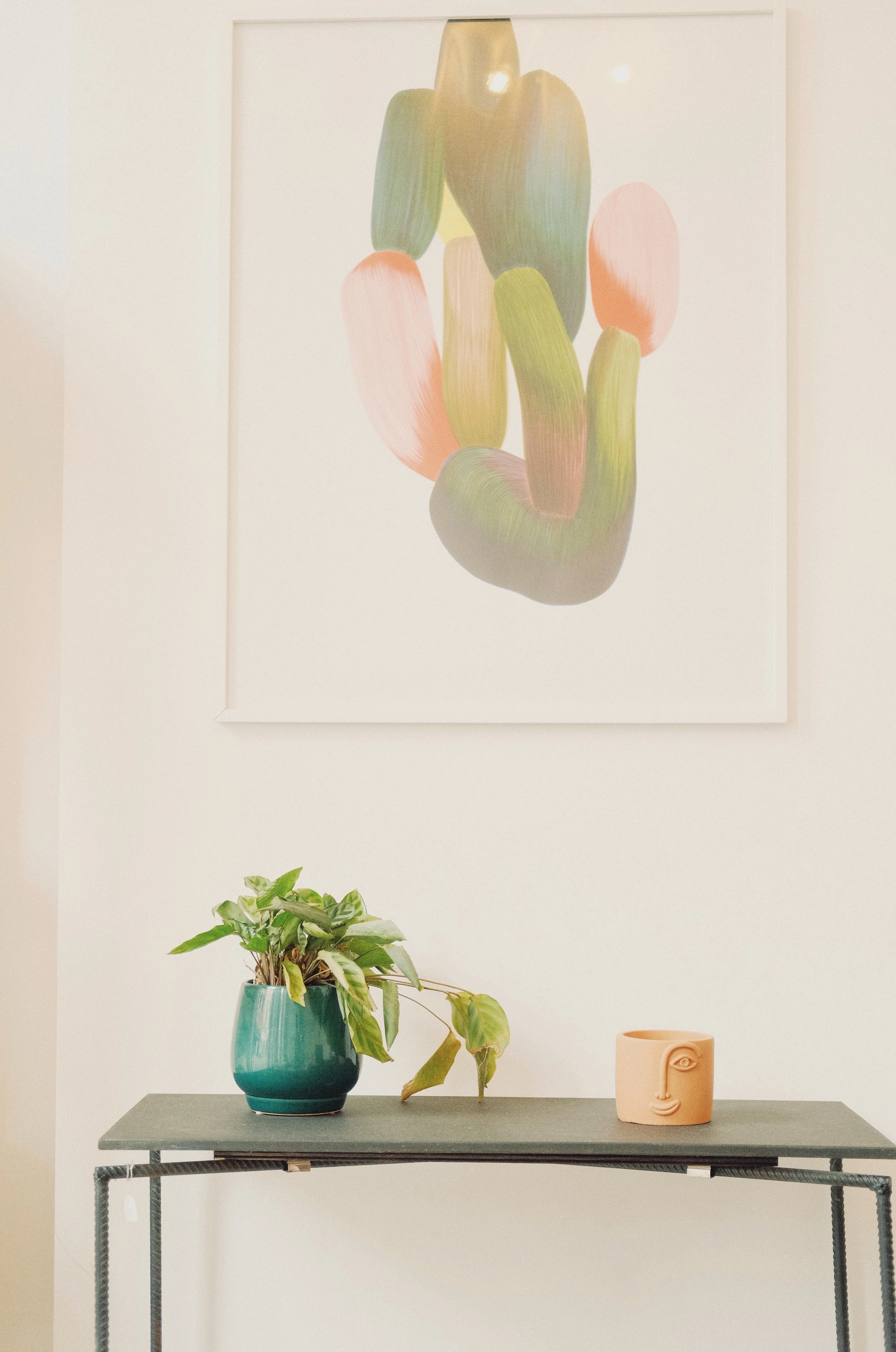 Une plante en pot vert, un objet en forme de visage humain en terre cuite, et une œuvre d'art abstraite représentant des formes organiques en vert, rose et beige sur le mur blanc. | QSphotography | Namur