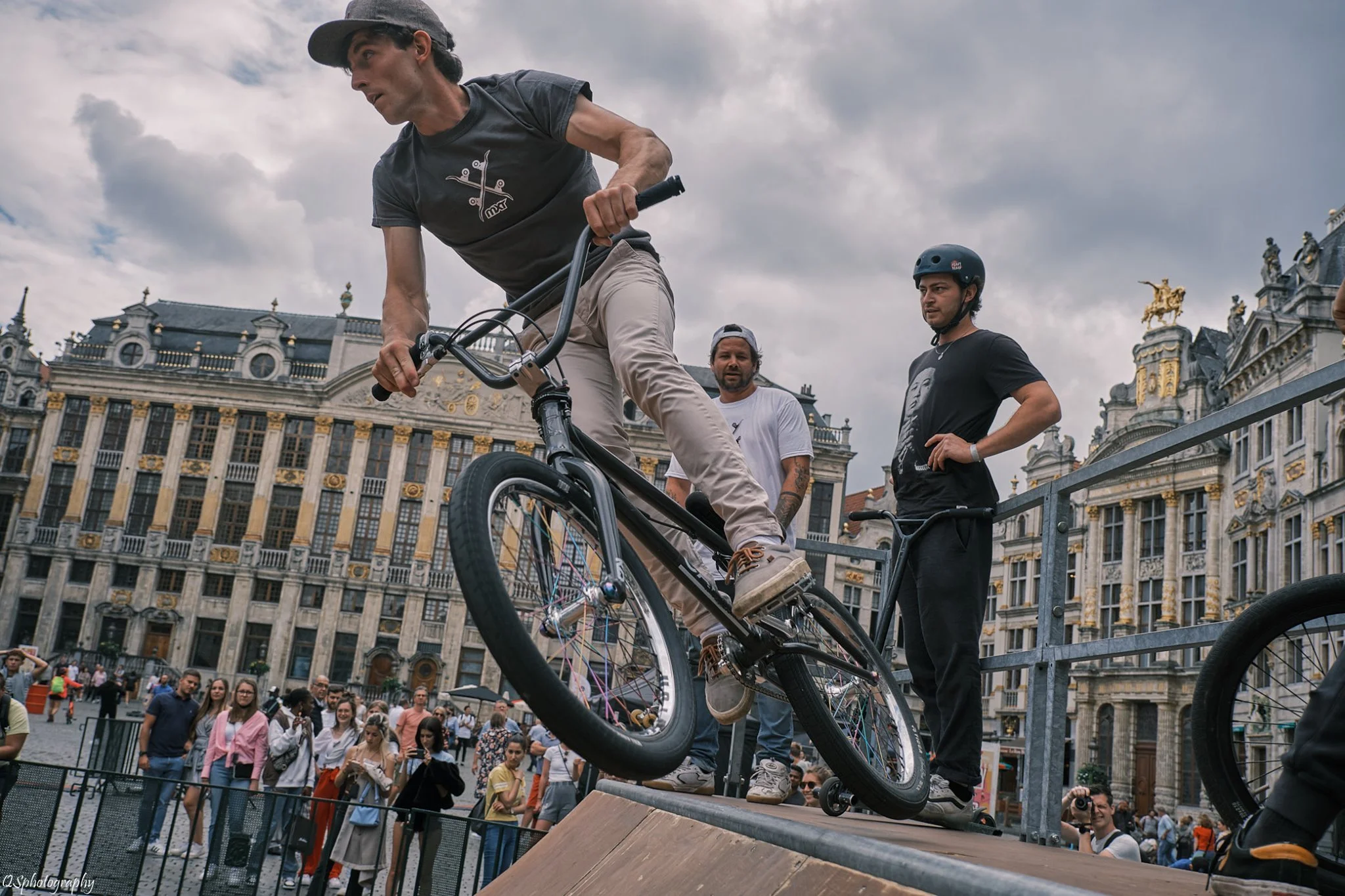 Philippe Cantenot effectue une figure sur un vélo dans une démonstration de skateboard ou BMX, avec un groupe de spectateurs et une architecture historique en arrière-plan. | QSphotography | Namur