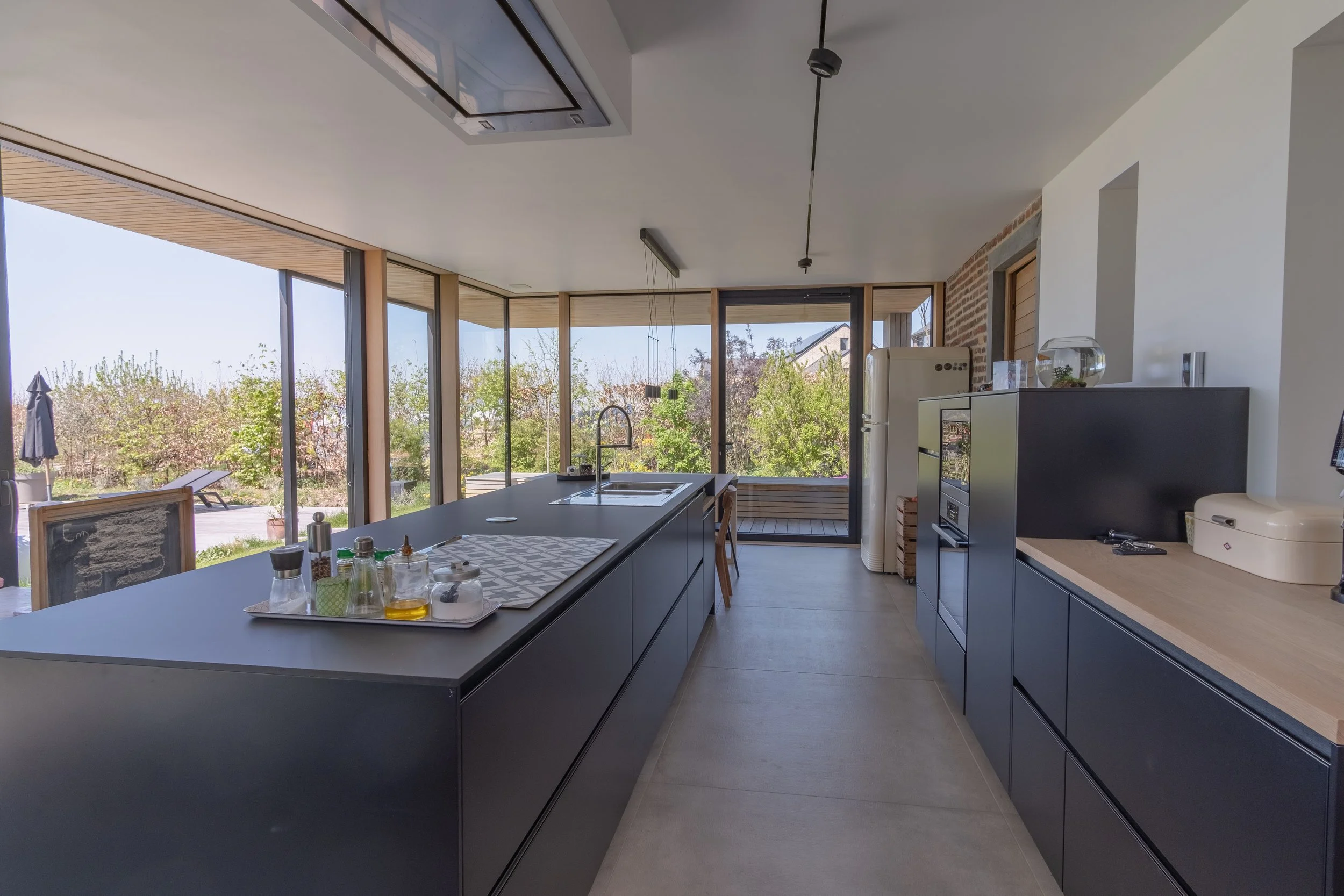 Cuisine moderne avec îlot central noir, fenêtres panoramiques, et vue sur un jardin, | QSphotography | Namur