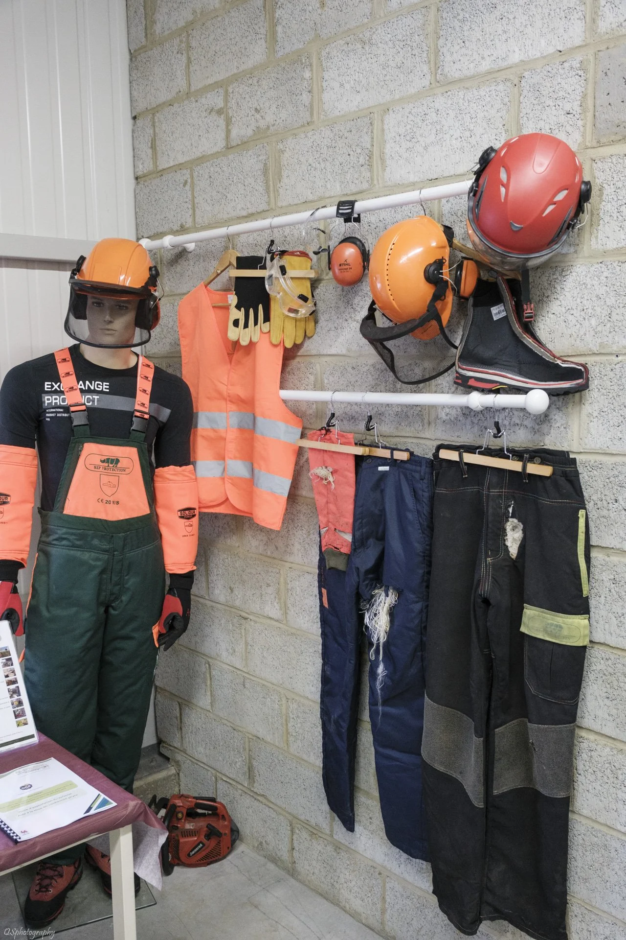 Équipement de sécurité et vêtements de travail suspendus et affichés sur un mur, y compris des casques, des gants, des lunettes de protection, des bottes, des vêtements de haute visibilité, des pantalons usés, et une mannequin portant un harnais de s