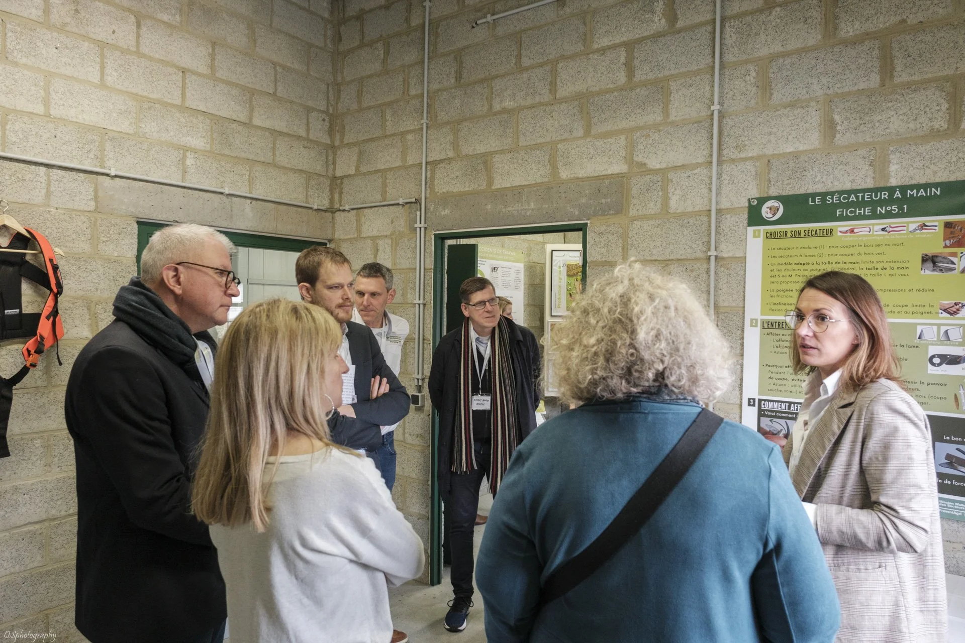 Groupe de personnes en train d’écouter une femme qui explique une affiche avec des instructions sur un processus dans une pièce avec murs en béton. | QSphotography | Namur