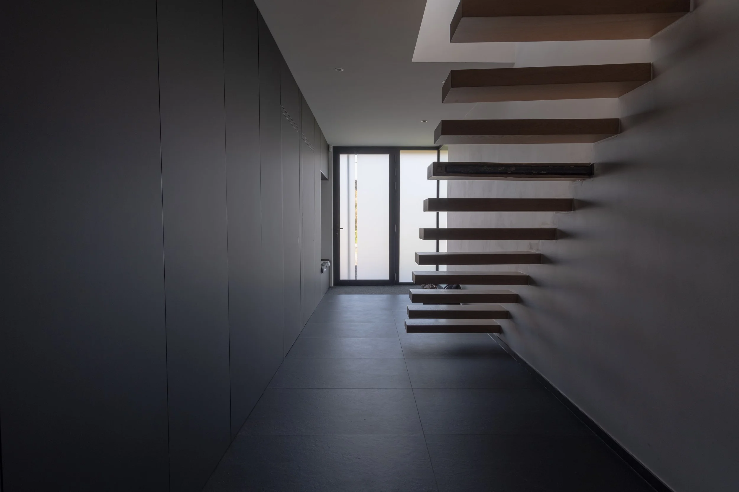 Hall d'entrée moderne avec porte vitrée, mur gris foncé, sol en carreaux noirs, et escaliers flottants en bois menant au-dessus. | QSphotography | Namur
