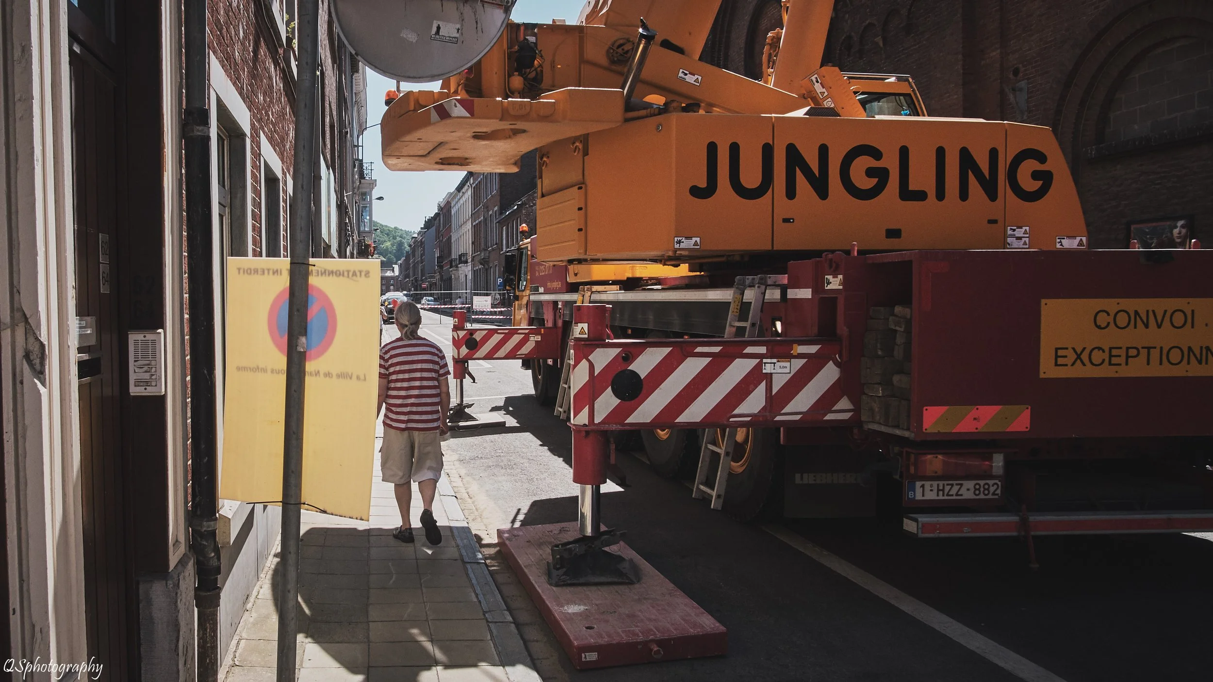Un homme marche sur le trottoir à côté d'un grand camion de travaux. Le camion est stationné en face d'un bâtiment ancien, et il y a un panneau de signalisation jaune avec un symbole interdit de stationnement. | QSphotography | Namur