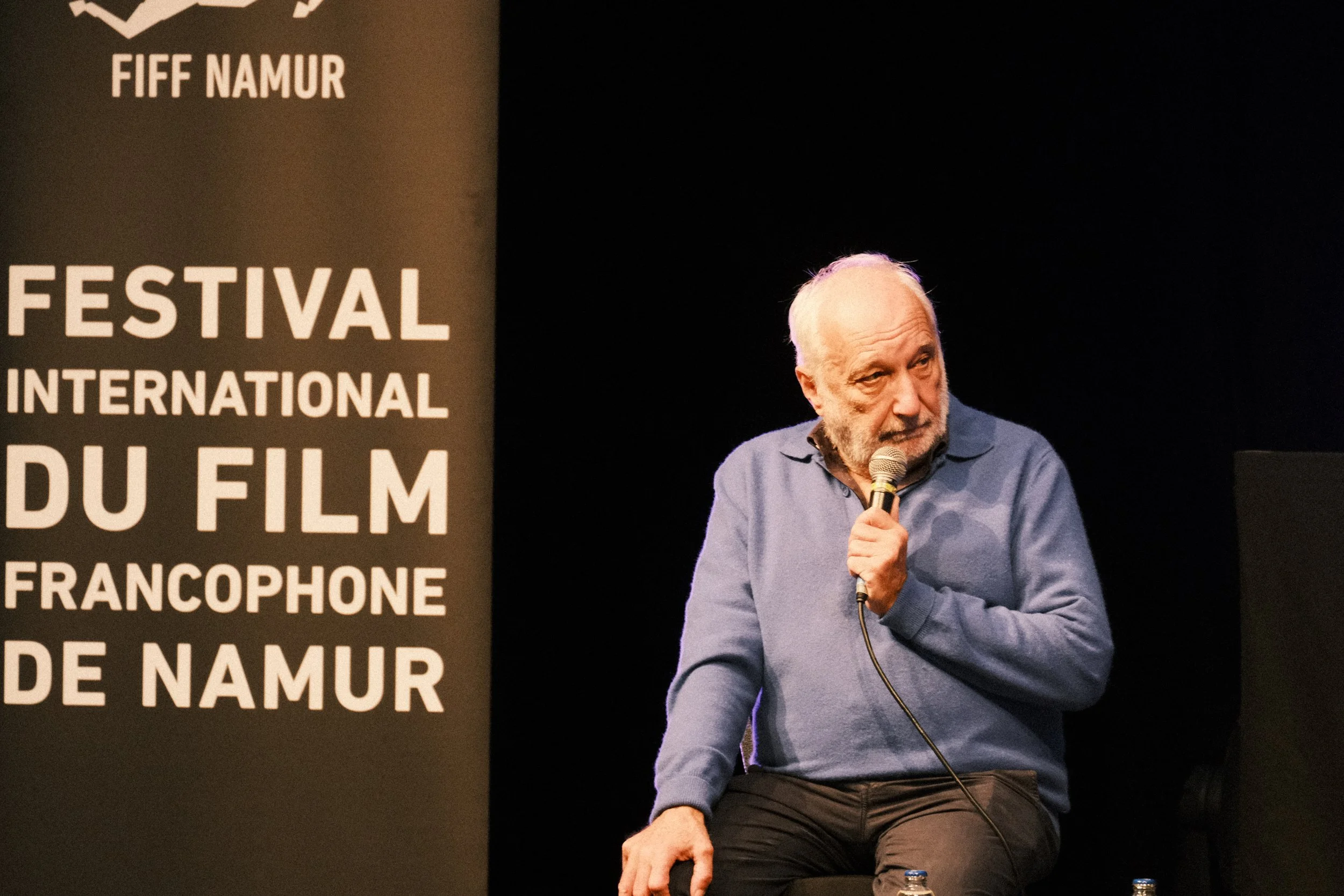 François Berléand portant un pull bleu, parle dans un microphone lors d'une conférence ou d'un événement, avec une bannière à gauche indiquant le Festival International du Film Francophone de Namur. | QSphotography | Namur