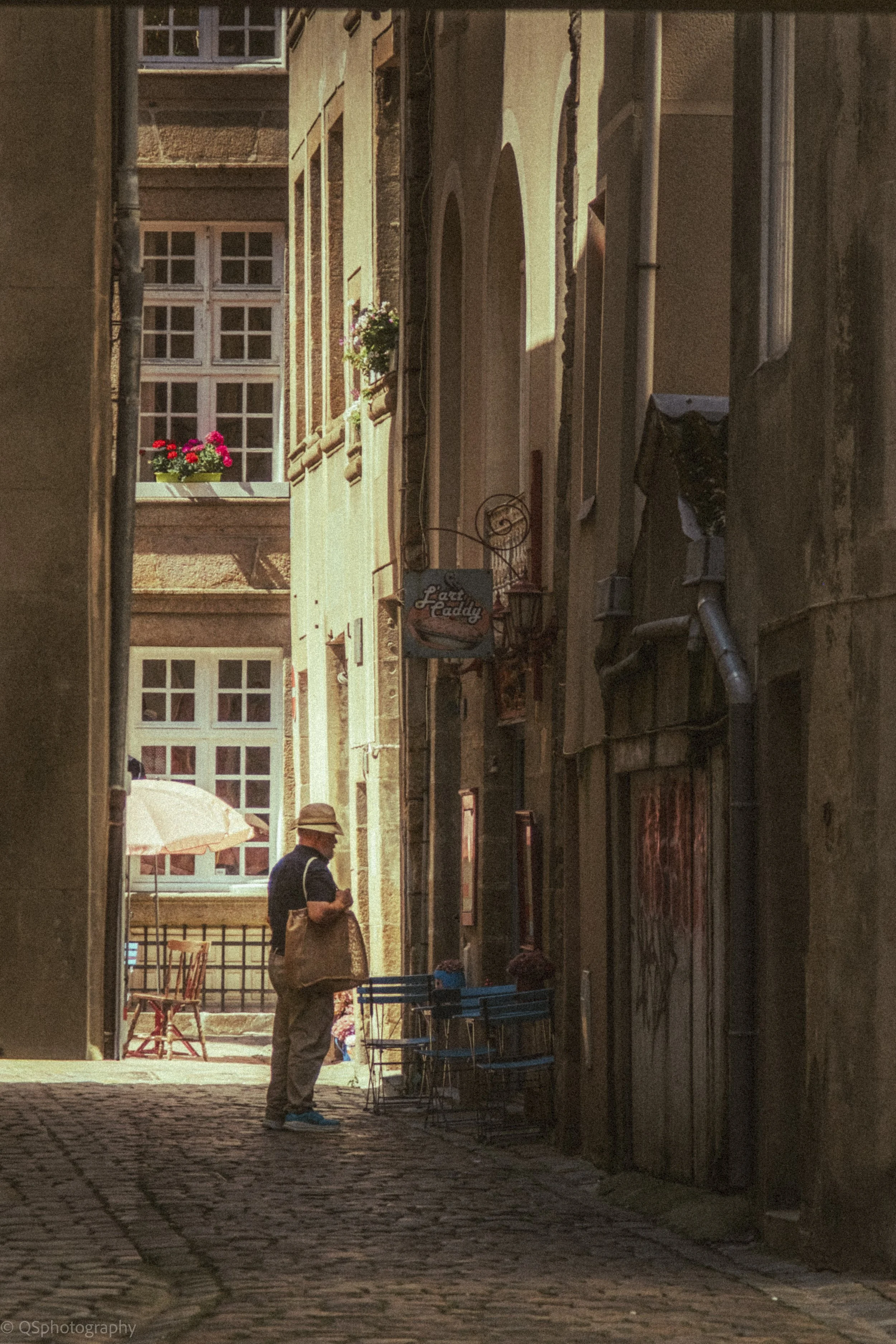 Une personne portant un chapeau, un t-shirt noir et un sac beige, se tient dans une ruelle pavée en Europe. Elle semble lire ou regarder quelque chose. La ruelle est bordée de bâtiments historiques avec fenêtres à carreaux et un panneau de signe de r