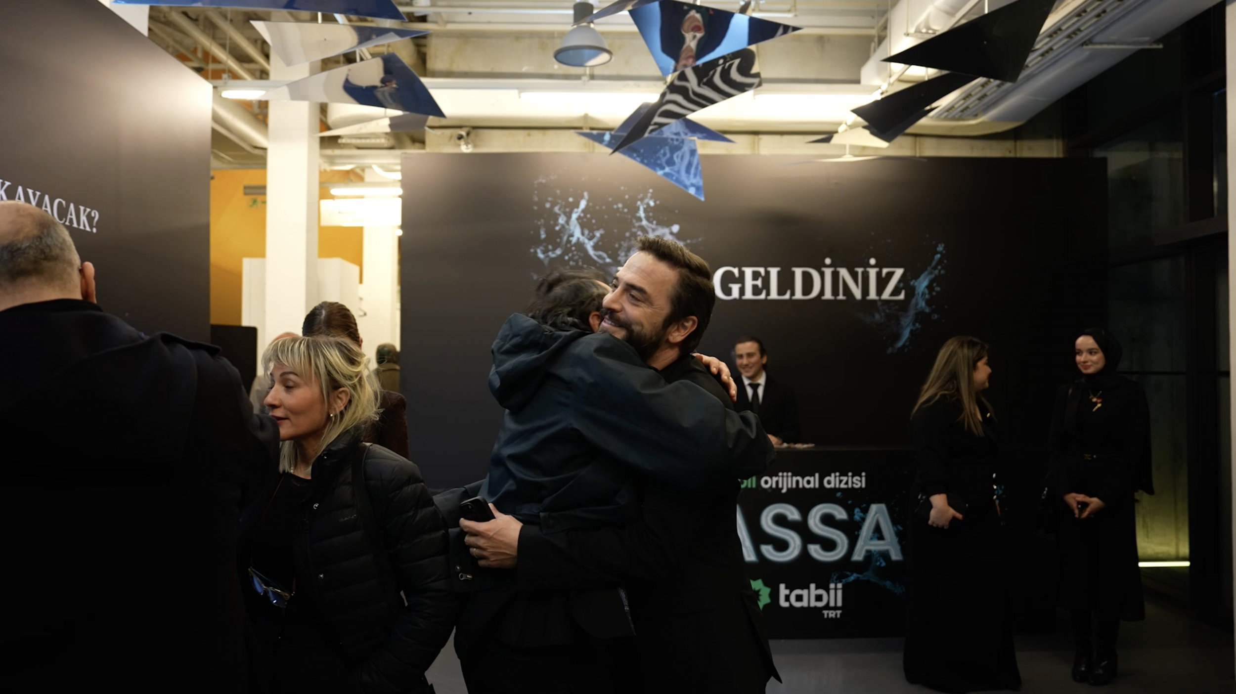 Two people hug warmly in the foreground at an event, smiling. Multiple other people are conversing and mingling in a modern indoor setting with a black backdrop displaying the words "HOŞGELDİNİZ" and promotional material for a TV series.