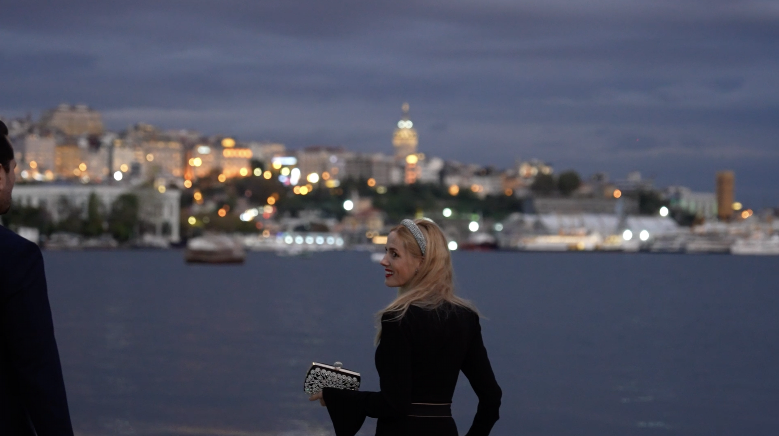 A woman with blonde hair, wearing a black dress and a gray headband, holding a clutch purse, smiling and looking to the right, with a blurred cityscape and harbor in the background during evening or night.
