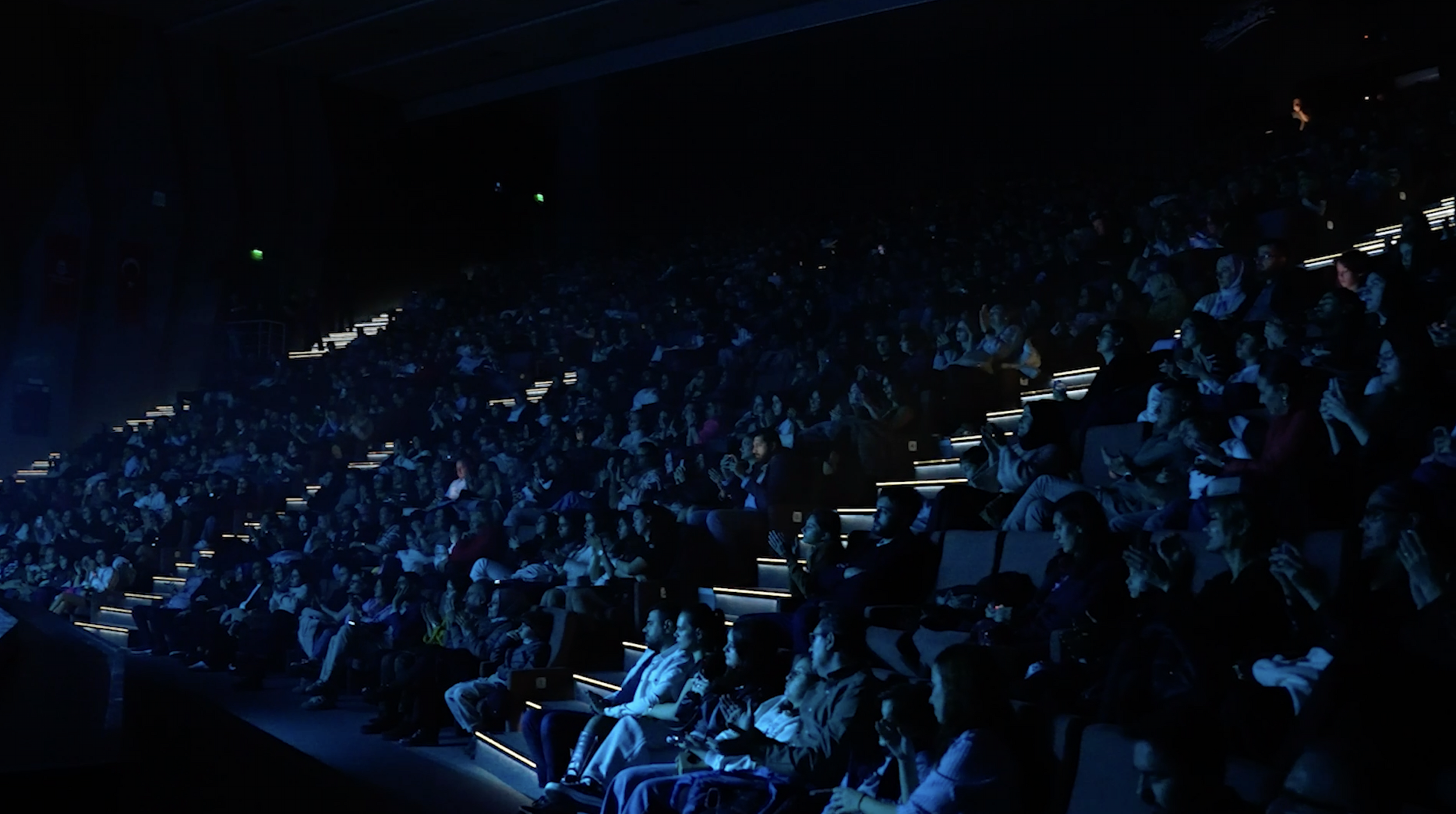 Audience seated in a dark theater or auditorium, some using electronic devices, with illuminated steps providing visibility.