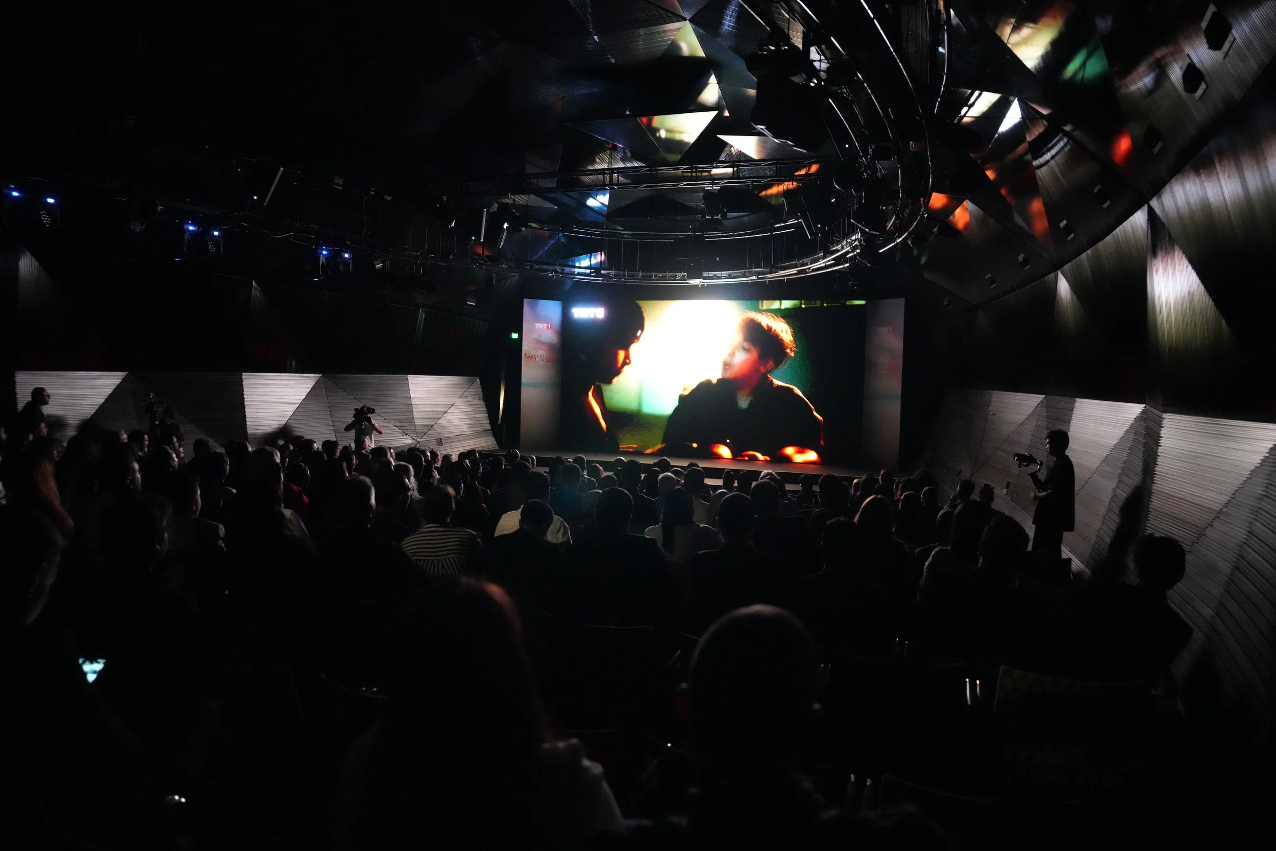 Interior of a cinema theater showing a movie on a large screen with audience seated in the dark.