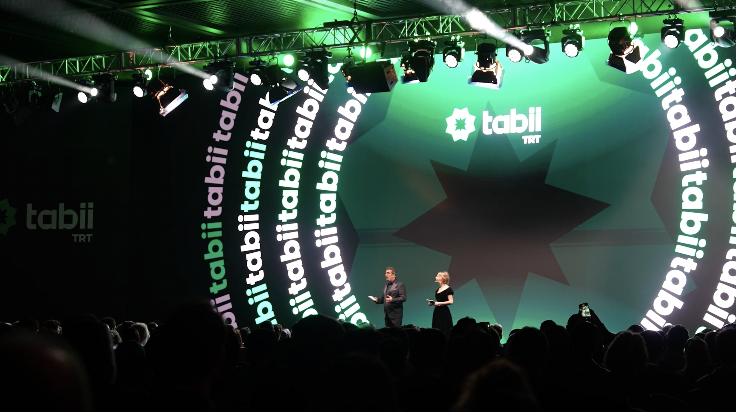 A stage with the backdrop displaying the logo and name 'tabii TRT' repeatedly in white and green text. There are lights and equipment hanging from above. Two presenters, a man and a woman, are standing on stage addressing an audience.