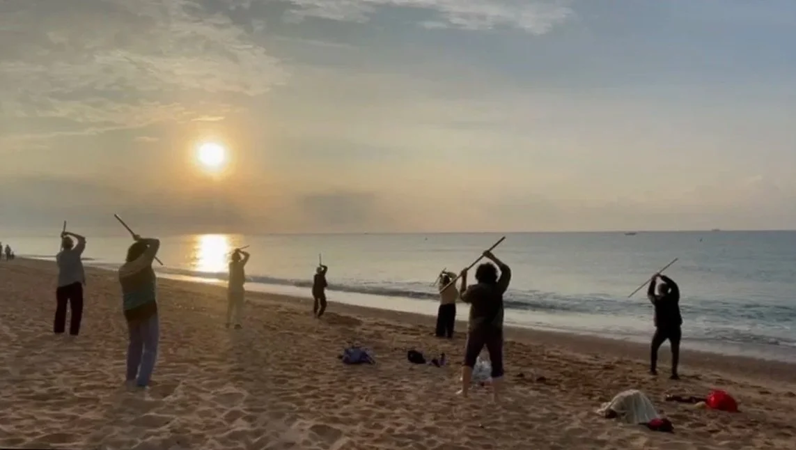 In this photo, 7 people are standing on the sand at the beach practicing Jo Kata, with a wooden Jo. They are facing a rising sun over the calm ocean.