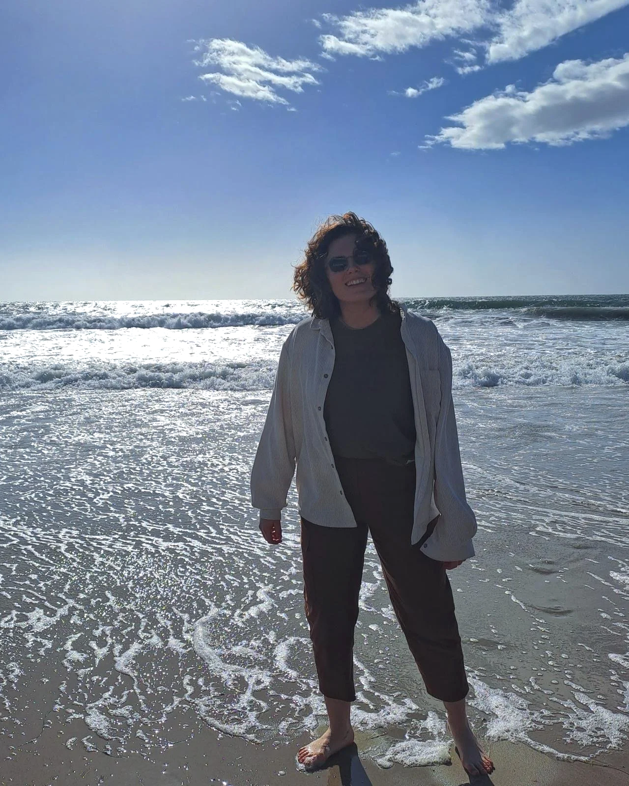 Clare is a white, nonbinary person who is standing at the ocean shore barefoot. They have mid- length curly hair, are wearing brown pants, grey tee and flannel. Behind them, there is a blue sky with some clouds and the ocean, with mid-sized waves.