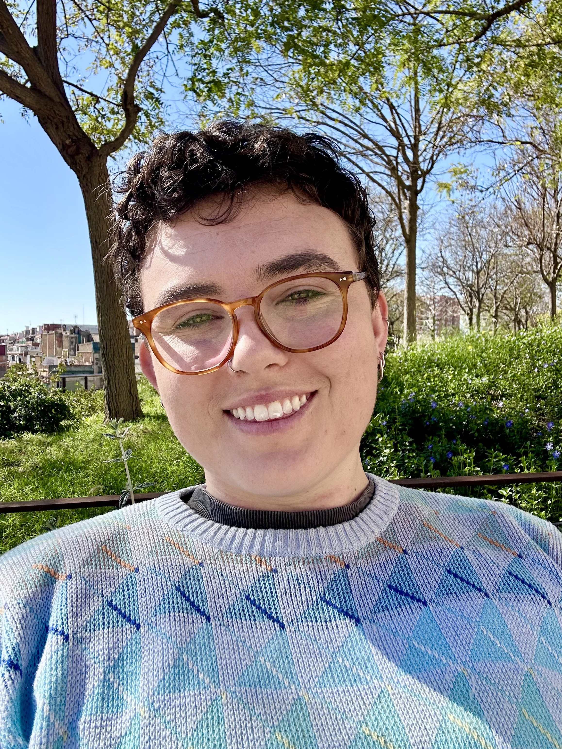 A photograph of Clare, a white nonbinary person with short, curly brown hair, dark eyebrows, and is smiling with teeth. They are wearing a blue sweater with a geometric pattern. In the background, there are green bushes, some purple flowers and trees