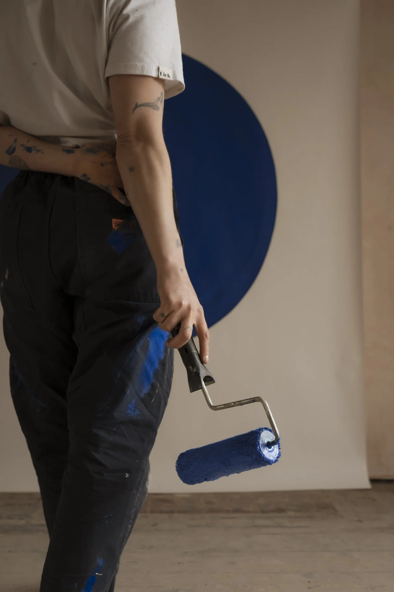 Person with tattoos holding a paint roller with blue paint, painting a wall in a room.