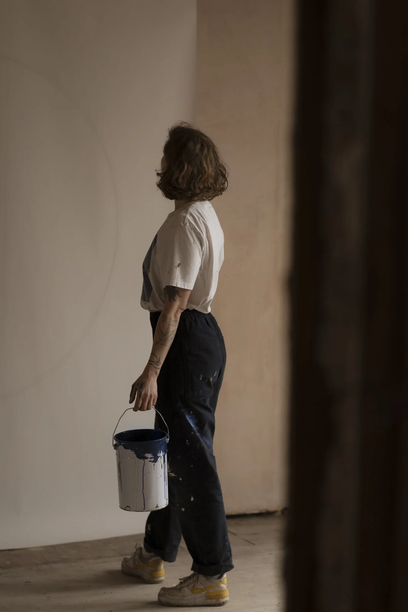 Person with curly hair wearing a white T-shirt and blue trousers, holding a paint bucket, standing on a wooden floor looking at a big paper sheet on the wall.