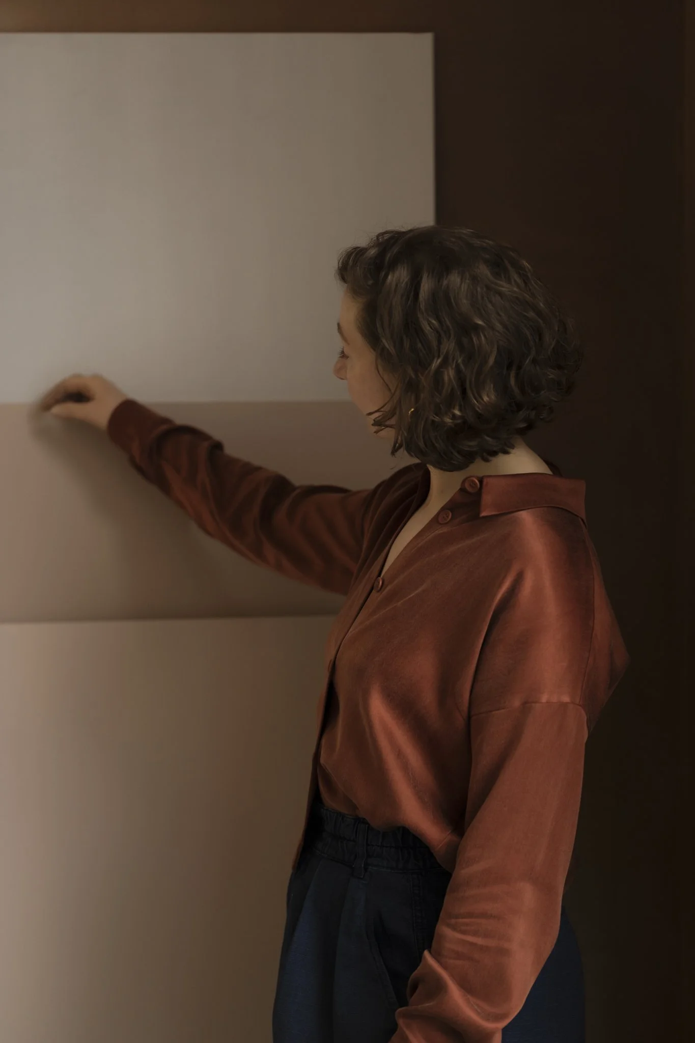 Woman with curly brown hair, wearing a silky satin bronze shirt and black pants, standing in front of big sheets of colour samples.