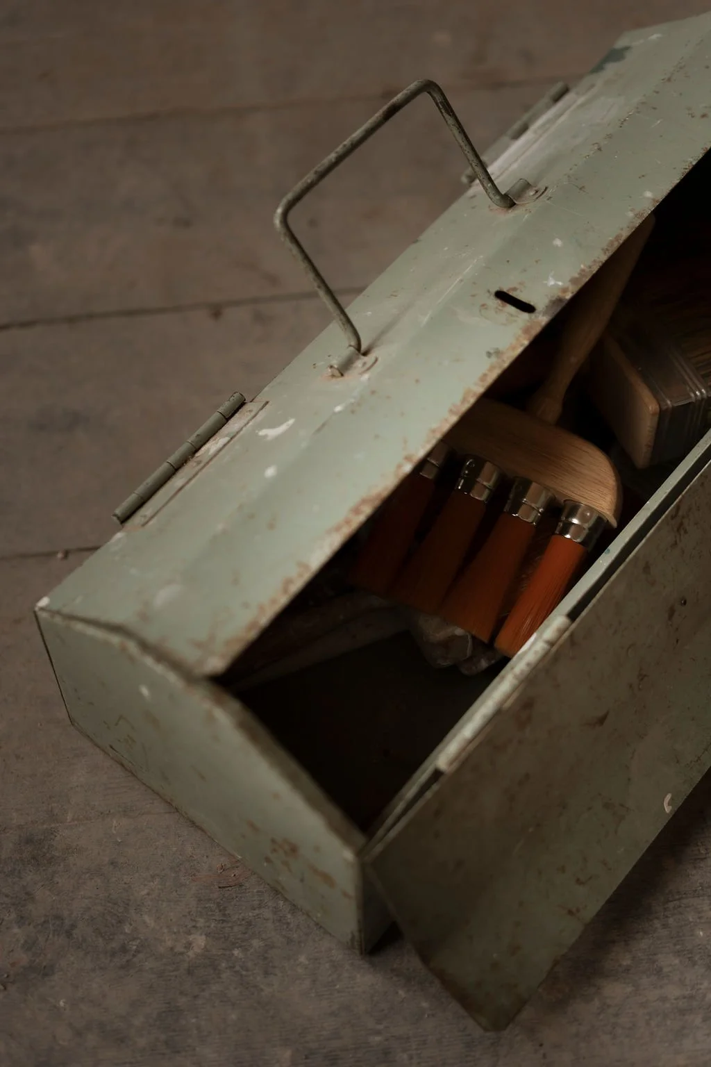 A rusty, old metal toolbox with an open lid, revealing a set of various paintbrushes inside.