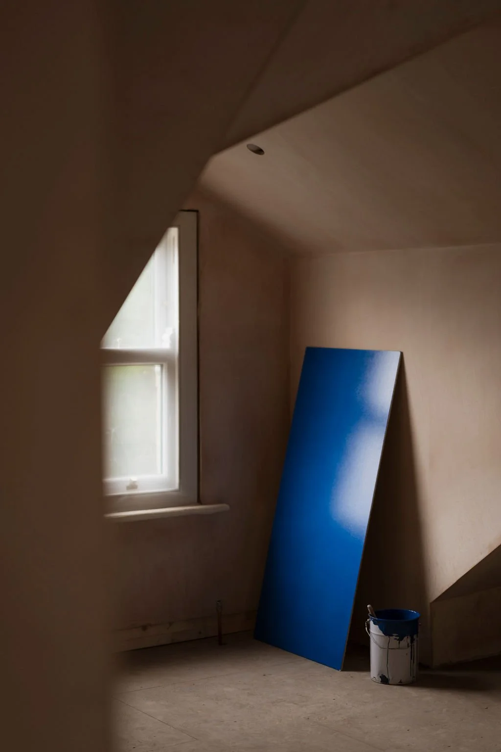 A blue painting leaning against a plastered wall near a window in a room under renovation, with a bucket of paint nearby.