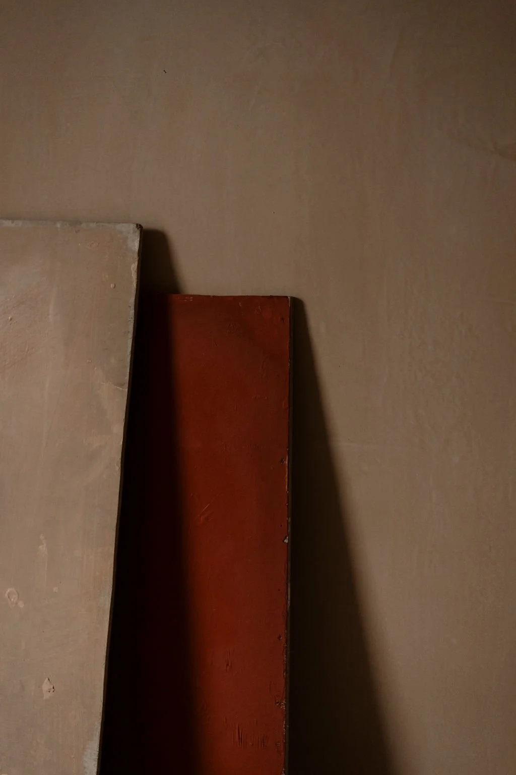 Close-up of two wooden boards, one painted red and the other light blush, leaning against a beige wall.