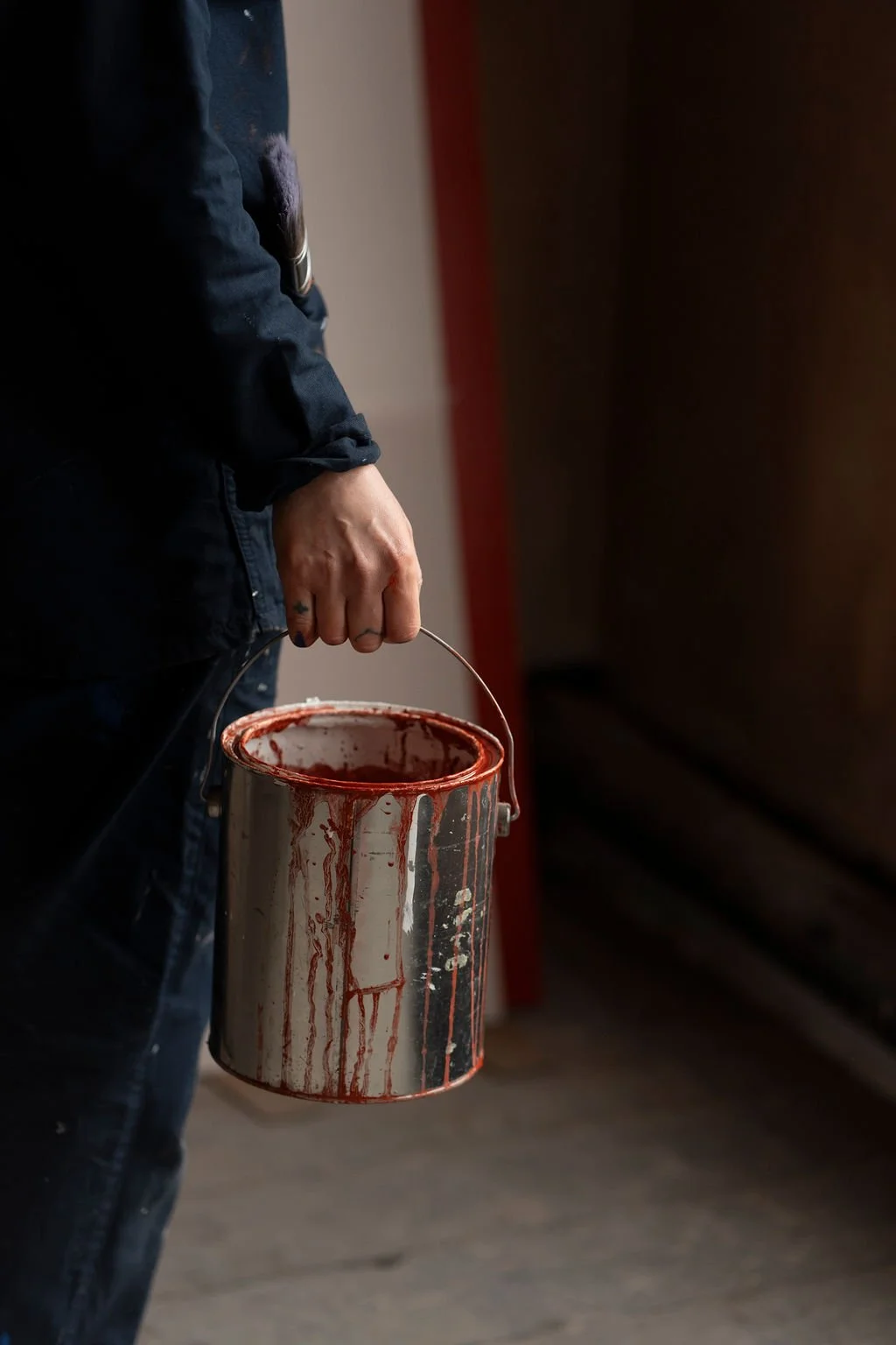 Person holding a paint can with red paint dripping down its sides, wearing a dark clothing.
