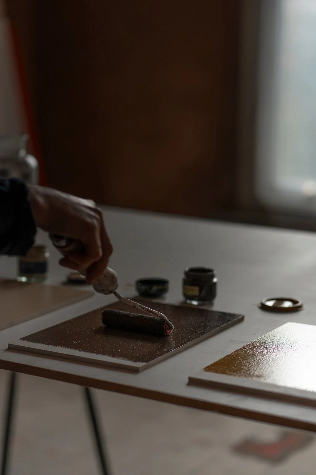 Person using an small paint roller in a dimly lit room with colour jars and tools around.