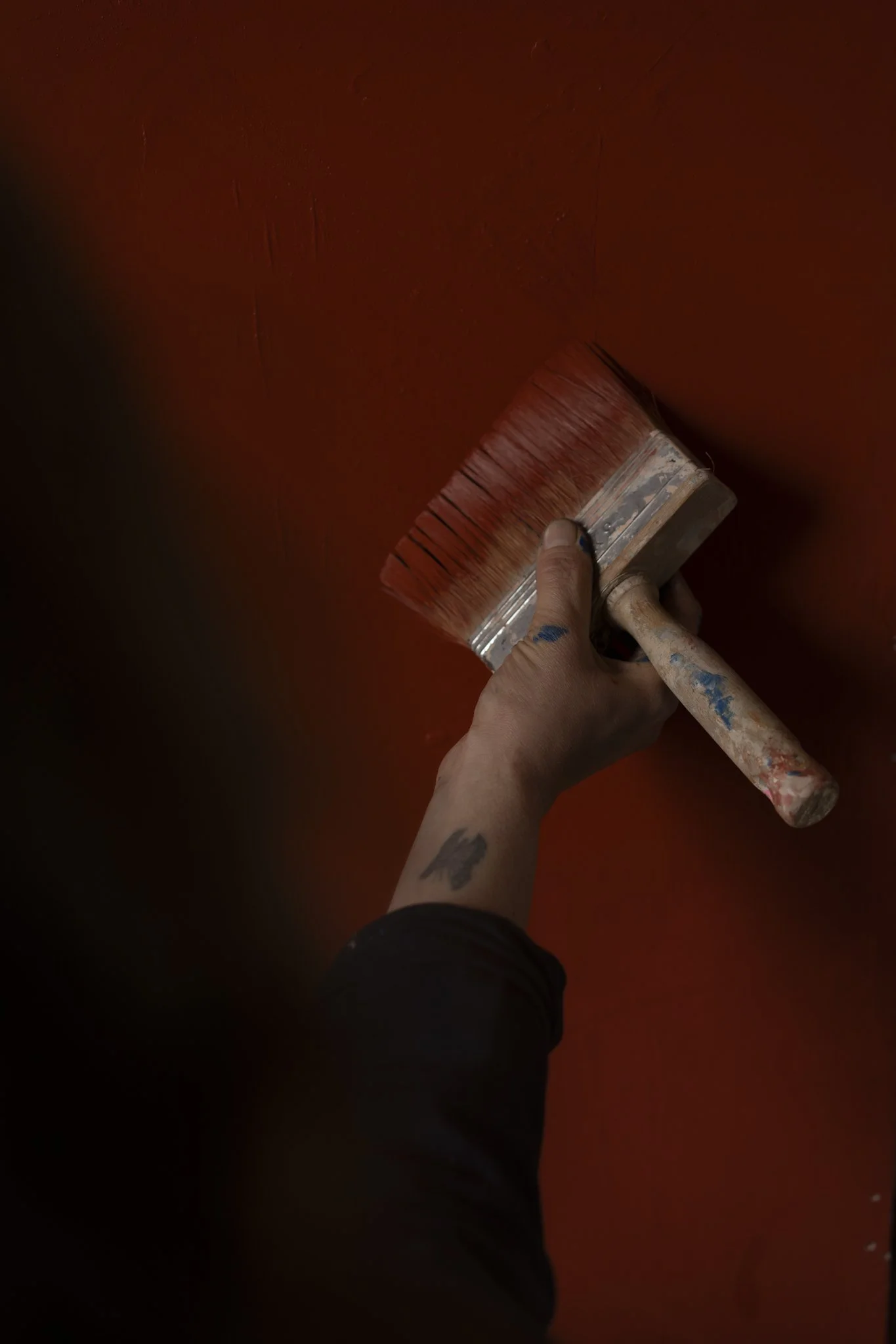 A person holding a limewash paintbrush and painting a wall red. The person's arm is visible, with a tattoo on the wrist, and they are wearing a dark sleeve.