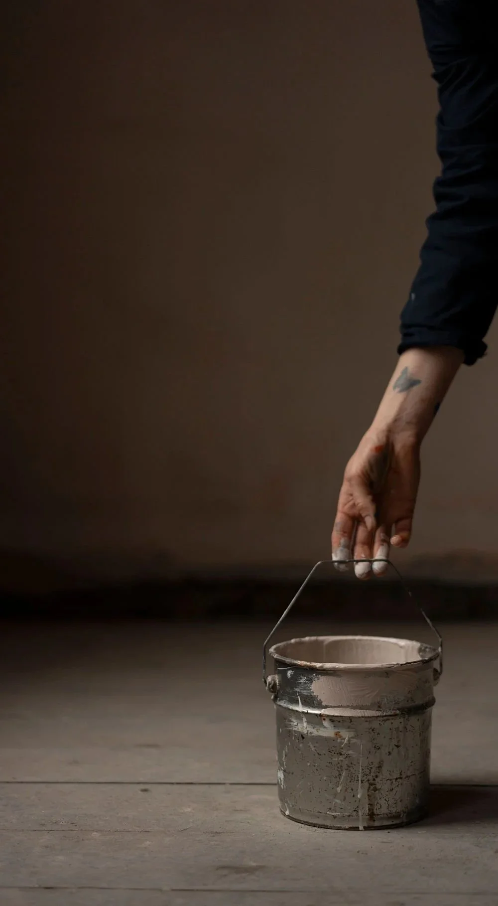 A person with tattoos on their arm is reaching into a paint bucket on a wooden floor, holding a paintbrush with blush paint on it.