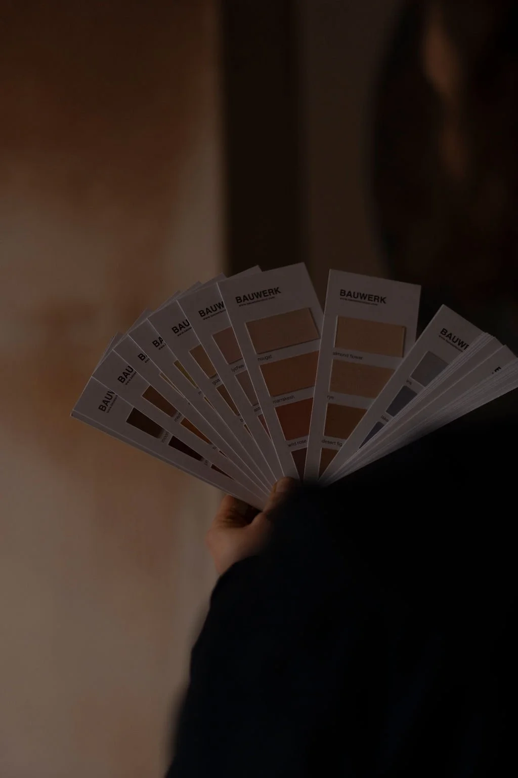 Person holding a fan of paint color swatches labeled Bauwerk, showing various shades of beige, brown, and grey.