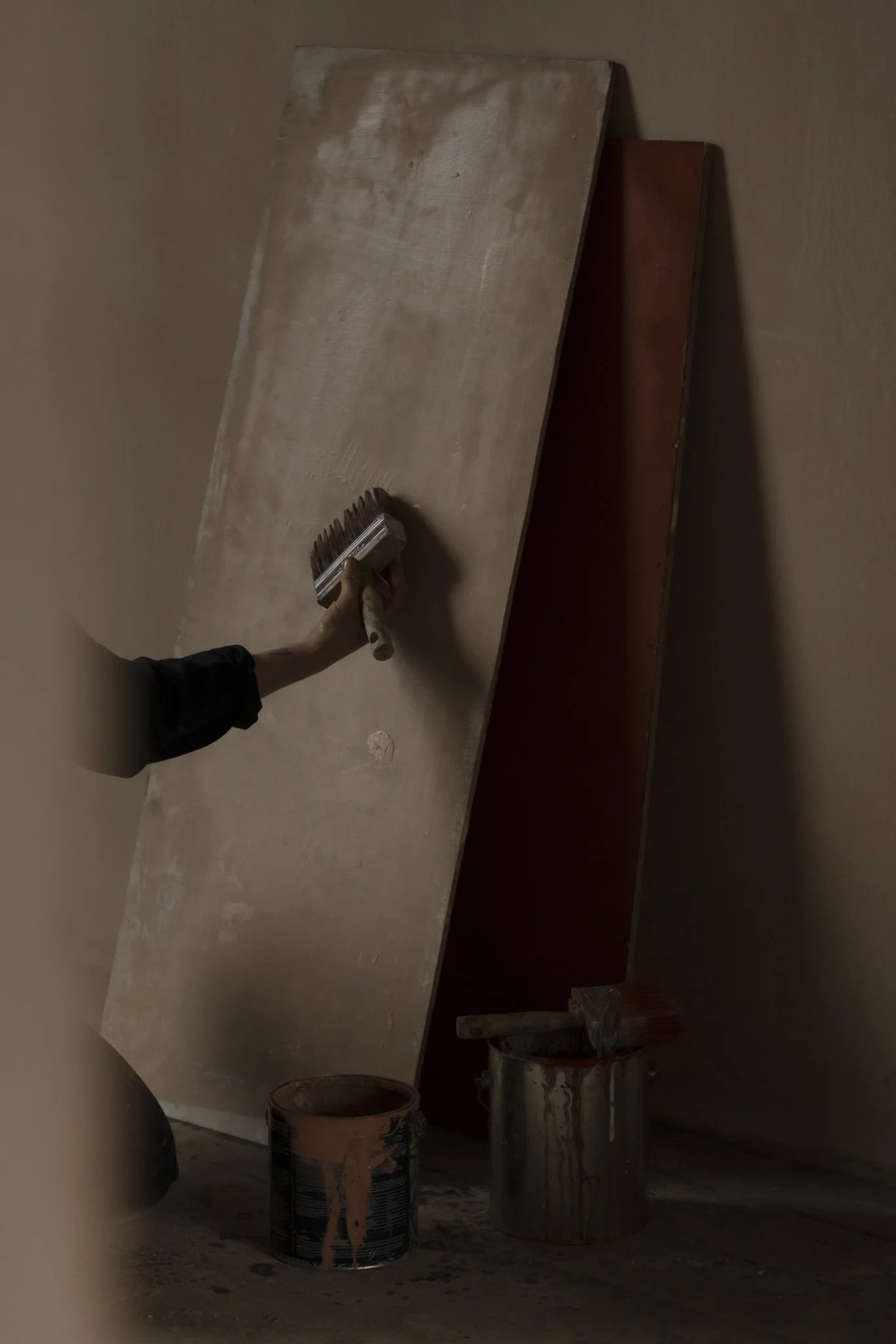 A person using a limwash brush to paint a wall with panel. Several paint cans, some with paint drips, are on the floor below the wall.