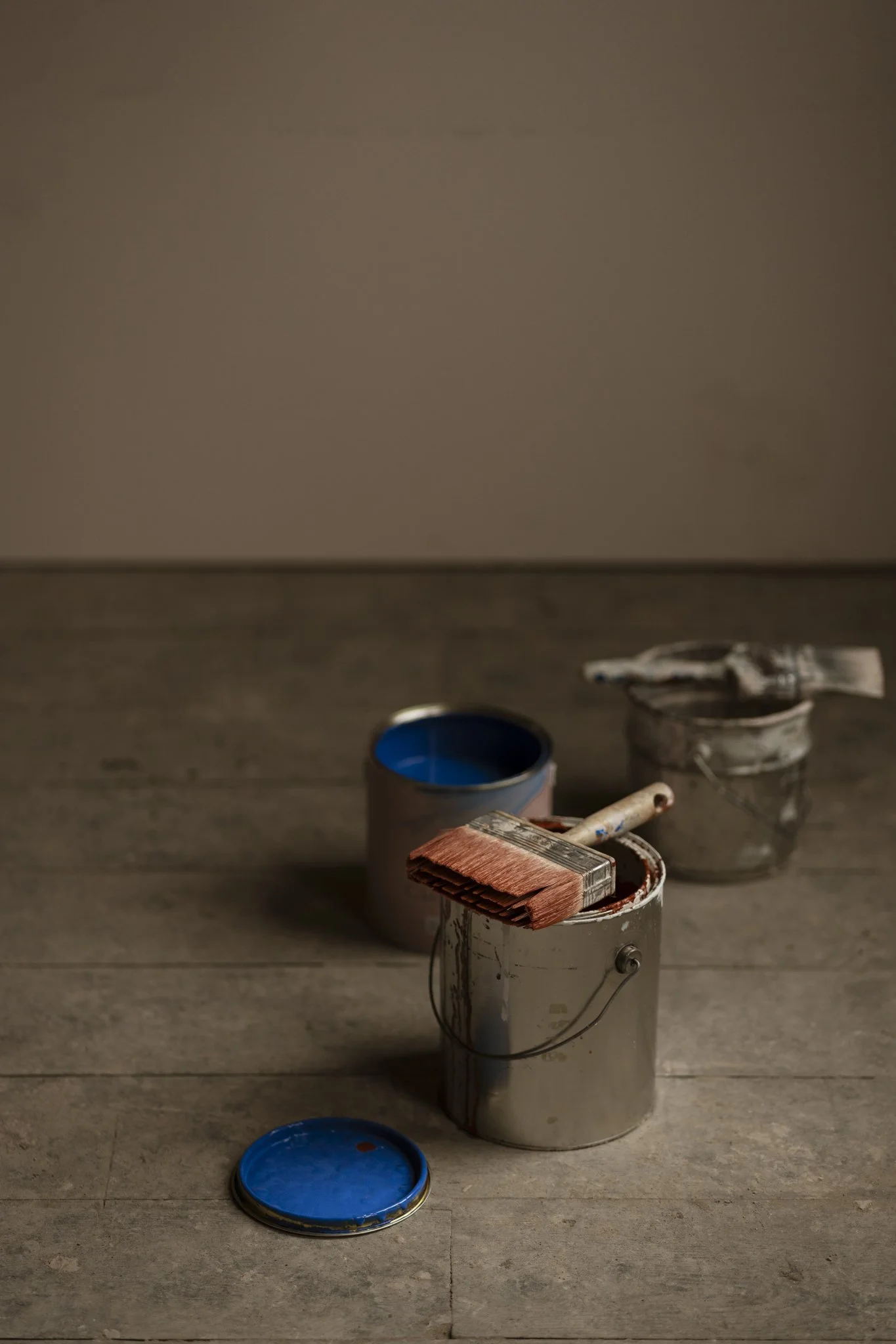 Several paint cans with brushes and lids on a wooden floor, some with dried paint marks, against a plastered wall.