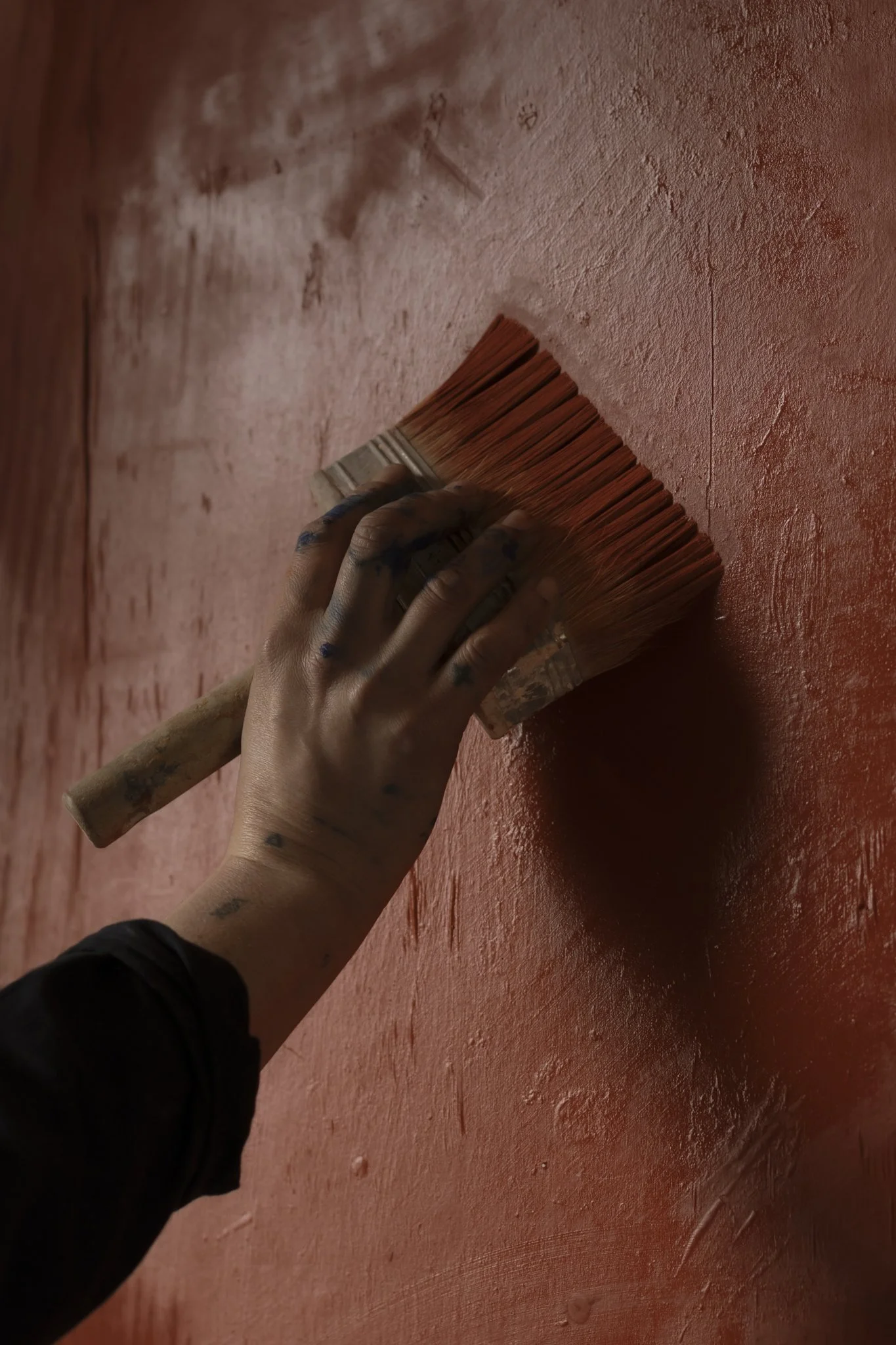 A person with tattooed hands using a limewash paintbrush to paint a wall in a reddish color.