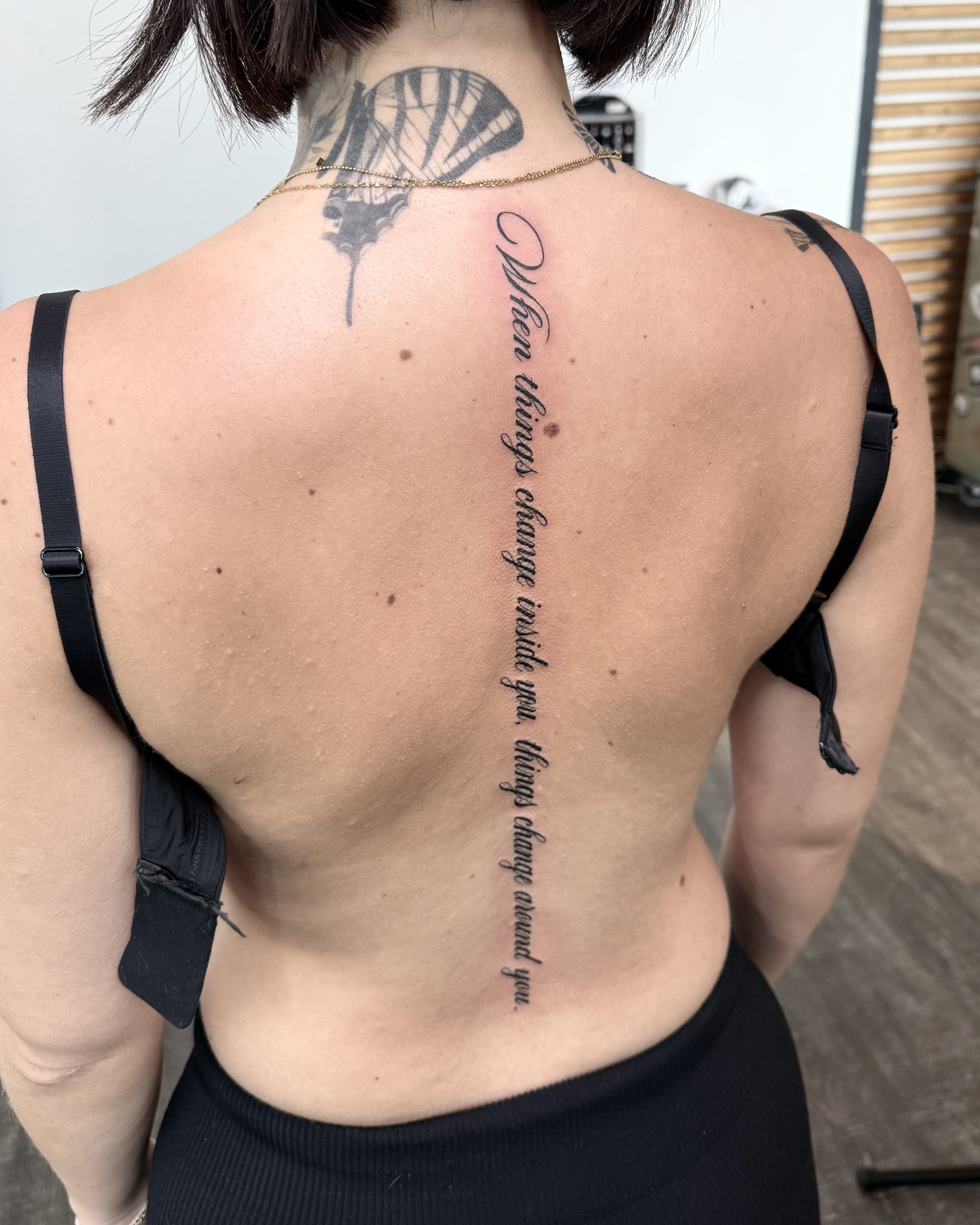 Rückenansicht einer Frau mit mehreren Tattoos, darunter ein Schriftzug in Französisch entlang der Wirbelsäule, ein Tattoo auf dem Nacken und ein schmetterlingsförmiges Tattoo auf dem Nacken.