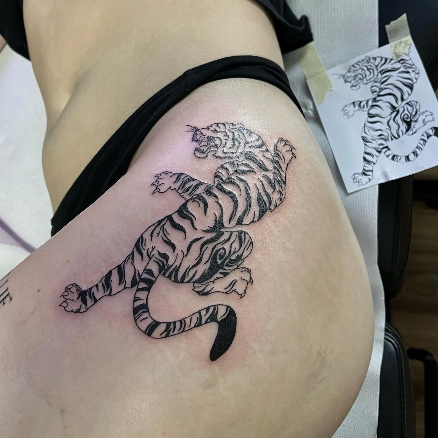 Tattoo eines Tigers auf dem Oberschenkel. Der Entwurf liegt auf Papier auf. Das Tattoo ist schwarz und grau mit Streifenmuster.