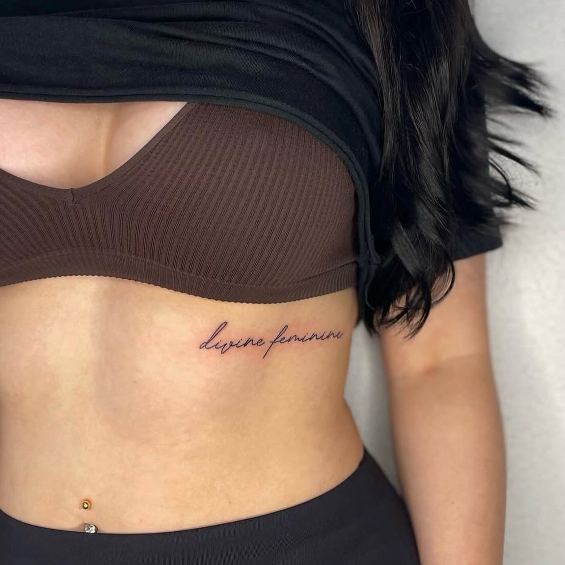 Nahaufnahme eines Tattoos auf der Seite eines Frauenkörpers, das den Schriftzug 'divine feminine' enthält, mit schwarzen Haaren und schwarzen Kleidung.
