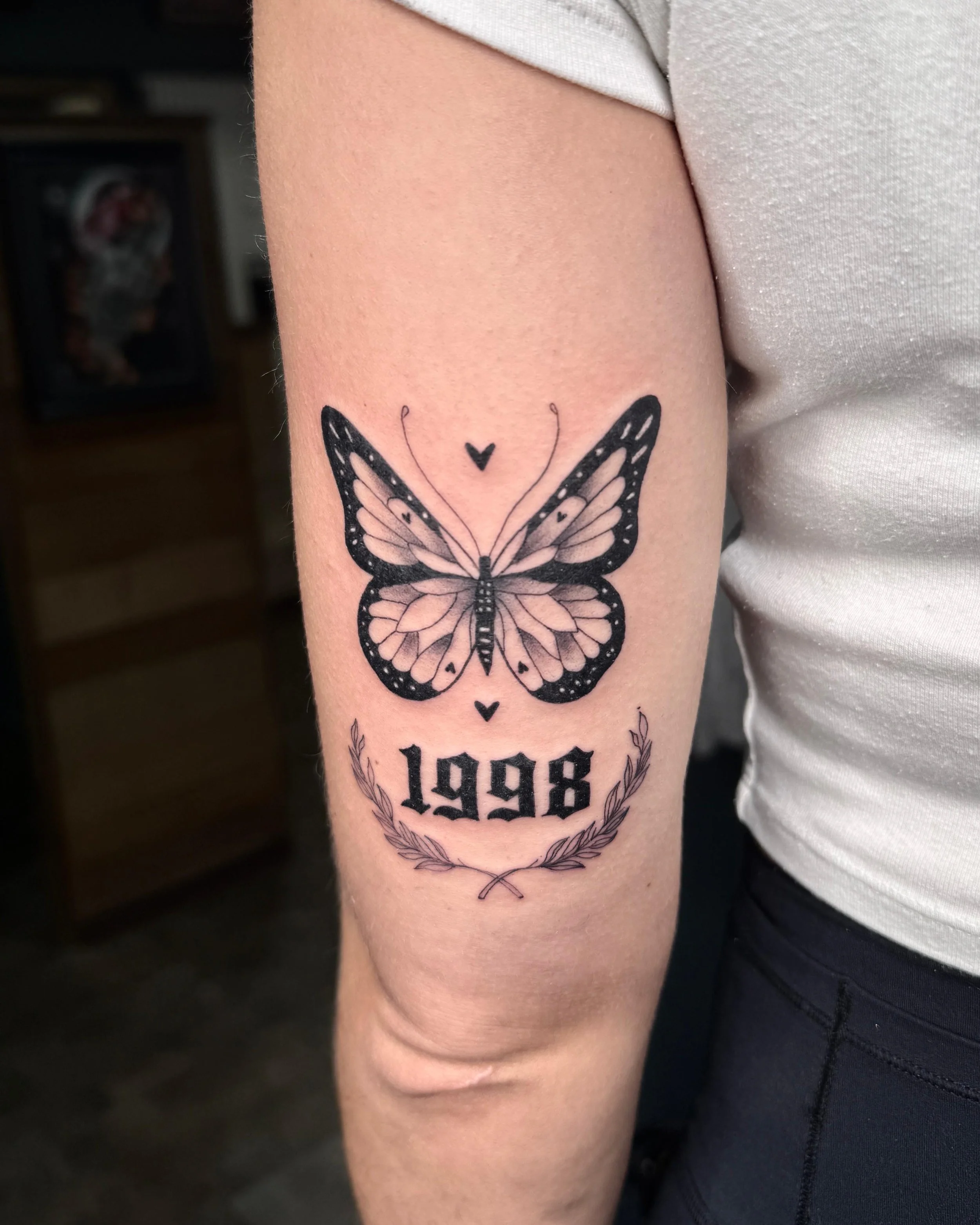 Tattoo eines Schmetterlings mit der Jahreszahl 1998 und einer Lorbeerkrone auf dem Oberarm.