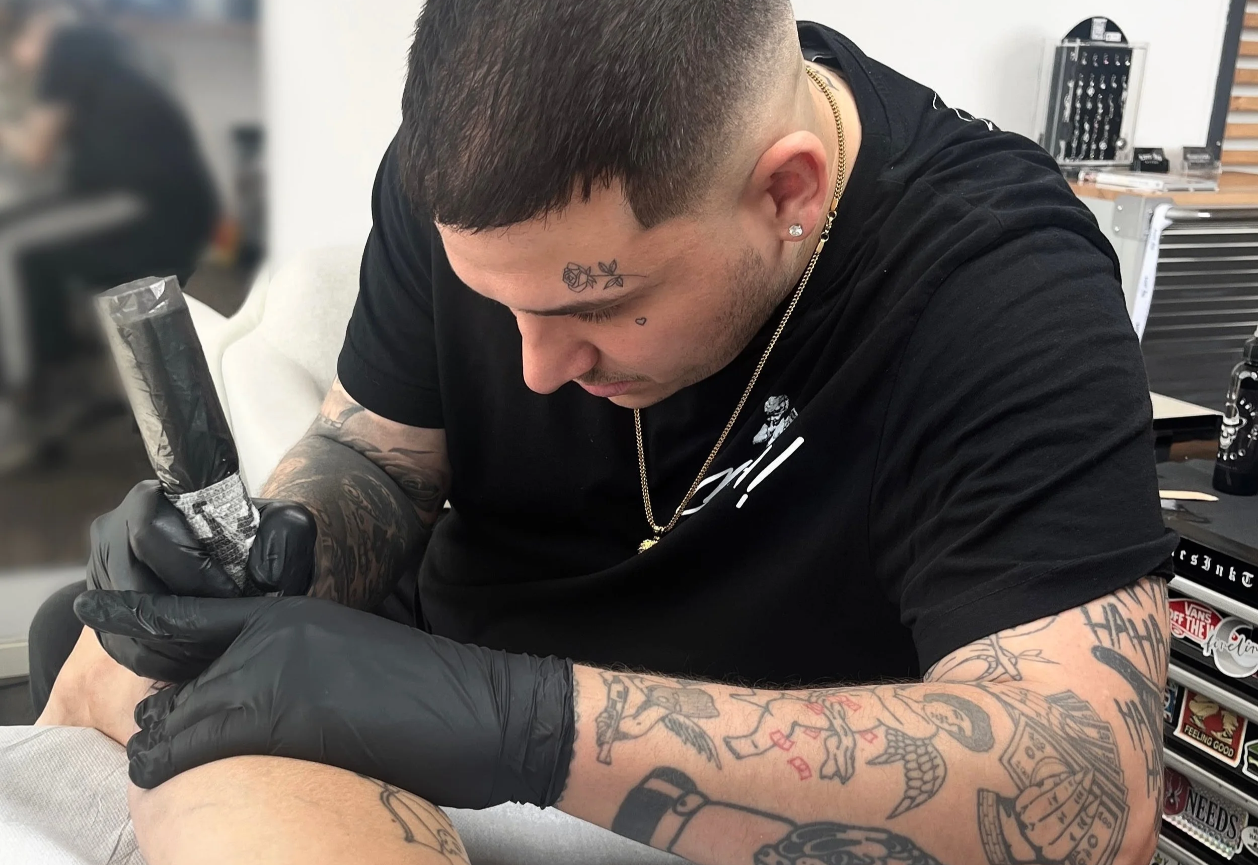 Tattoo-Künstler tätowiert eine Frau mit dunklen Haaren und Tattoos am Arm in einem Tattoo-Studio.