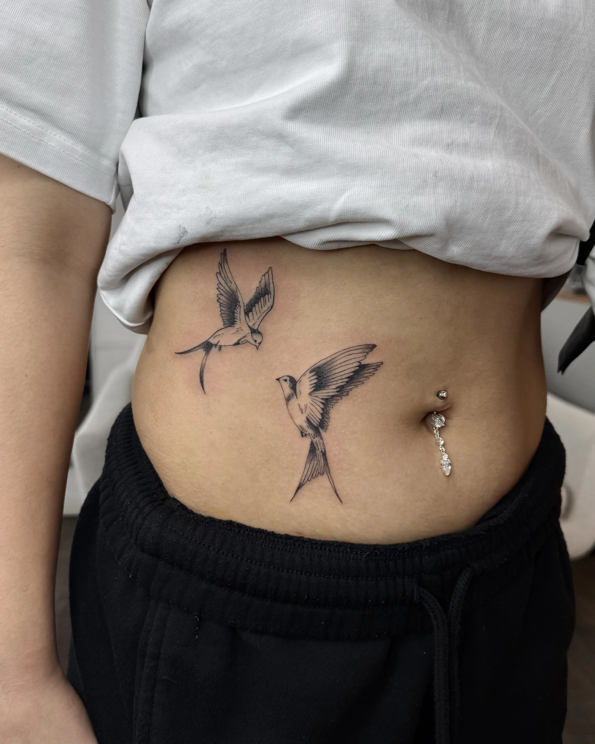 Tattoo von zwei fliegenden Vögeln auf dem Bauch, mit Nabelpiercing und schwarzem Hosenbund