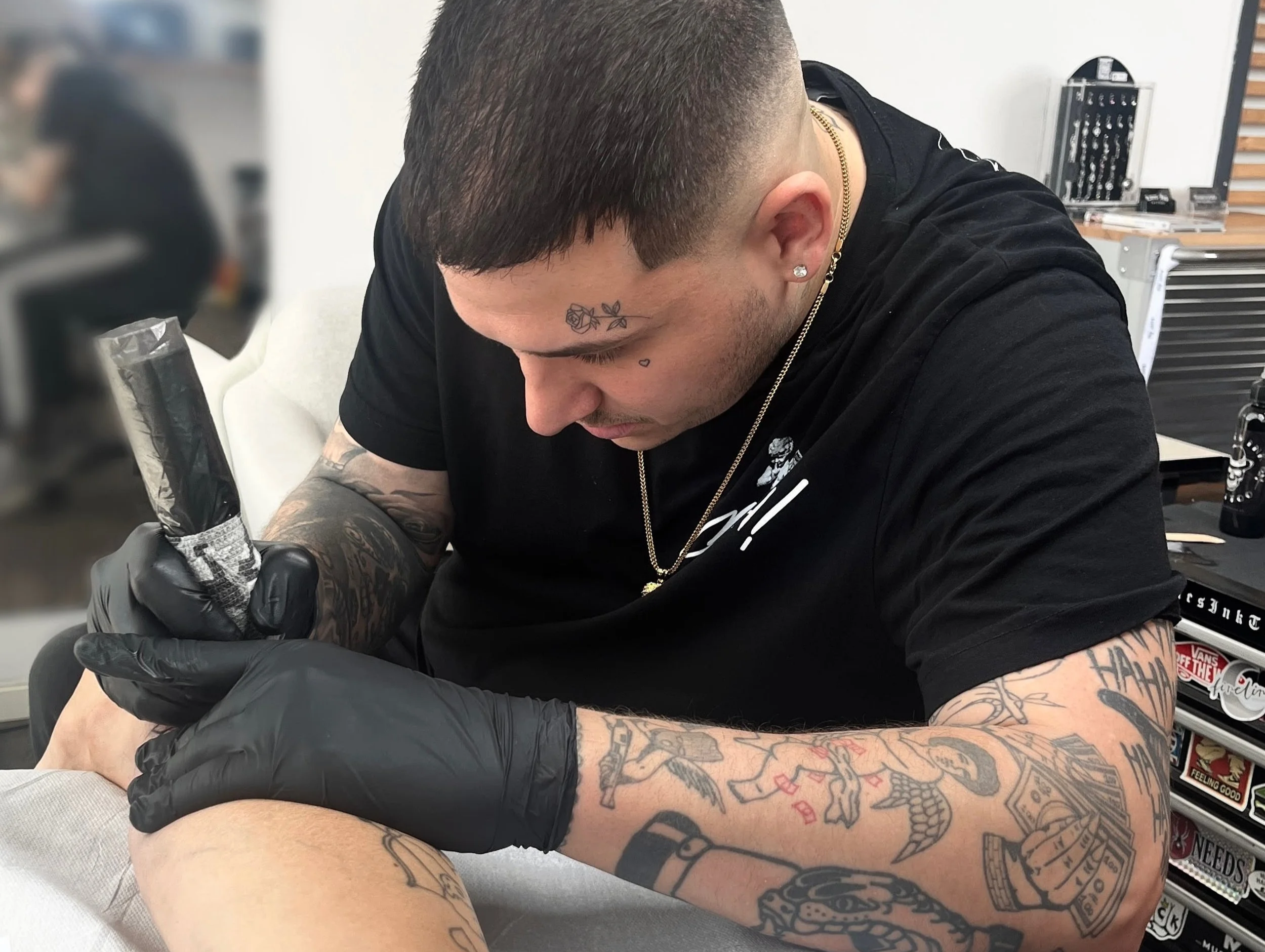 Tattoo-Künstler tätowiert einen Oberschenkel, trägt schwarze Handschuhe und einen schwarzen T-Shirt in einem Tattoo-Studio.