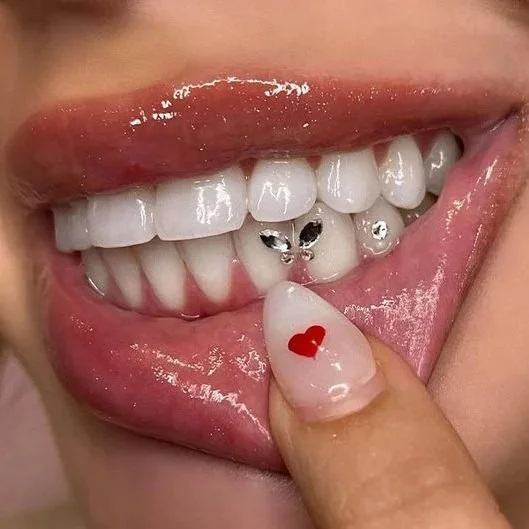 Nahaufnahme eines lachenden Mundes mit Zähnen, die mit Piercings und kleinen Schmuckstücken verziert sind, und einer Person mit einer Nagelfeile, auf der ein rotes Herz gemalt ist.