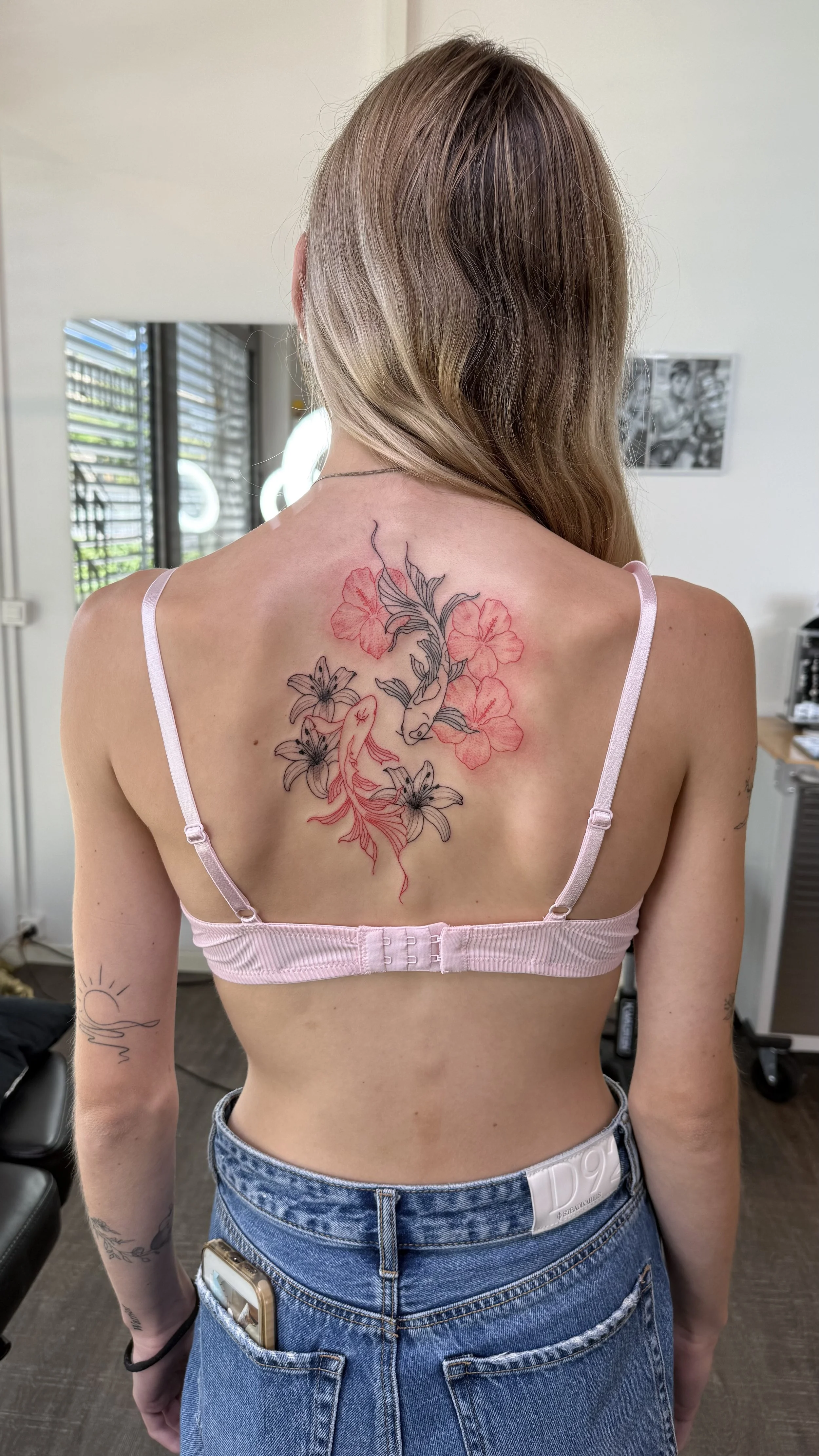 Eine Frau mit langen braunen Haaren trägt ein pinkes Top und eine Jeanshose. Auf ihrem Rücken ist ein farbiger Tattoo eines Fisches und Blumen sichtbar.