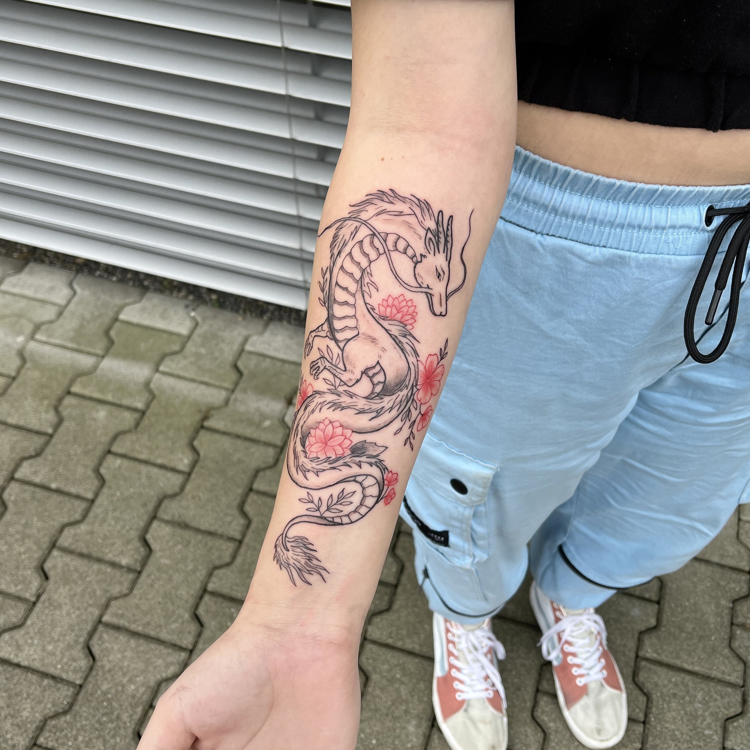 Tattoo eines Drachen mit roten Blumen auf dem Unterarm einer Person.