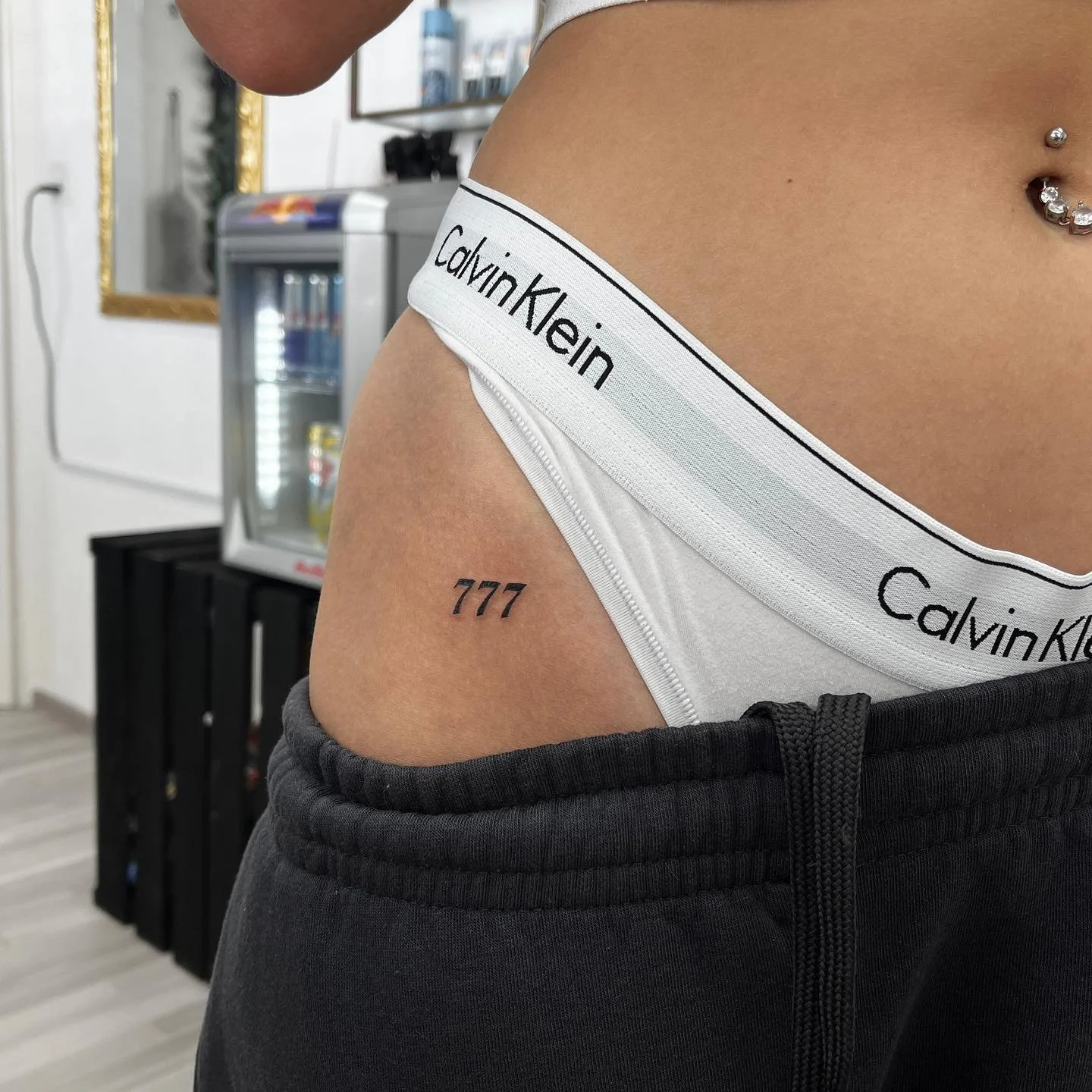 Nahaufnahme einer Person mit Tattoo '777' am linken oberen Oberschenkel, die weiße Calvin Klein Unterwäsche trägt, die Kleidung sitzt auf der Hüfte, im Hintergrund sind ein Wasserautomat und ein Spiegel sichtbar.