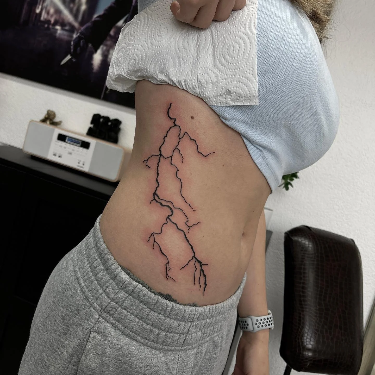 Tattoo eines Blitzes auf der rechten Seite des Bauchs einer Person, die eine graue Hose und ein weißes Oberteil trägt.