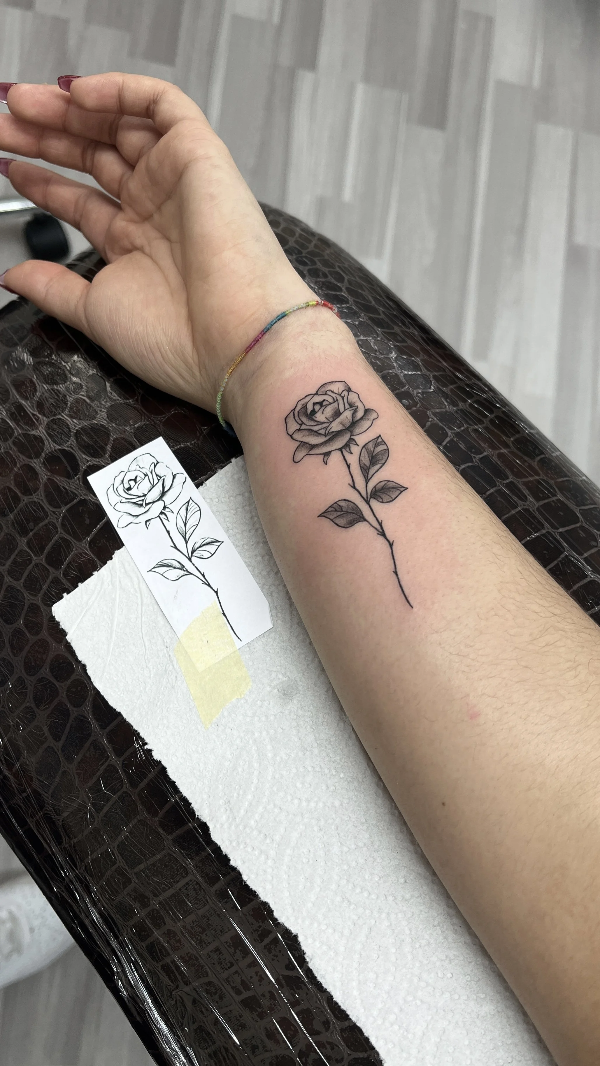 Hand mit einem schwarzen und grauen Rosen-Tattoo am Unterarm, daneben ein Tattoo-Vorlage und einen Tupfer.