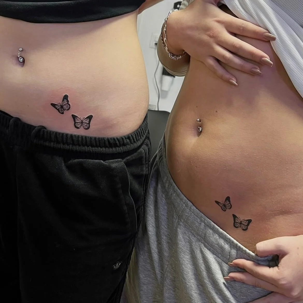Zwei Frauen mit Tattoos und Bauchnabelpiercings, eine trägt schwarze Shorts, die andere trägt graue Shorts, beide halten ihre Oberkörper frei, Blick auf ihre Bäuche und Tattoos.