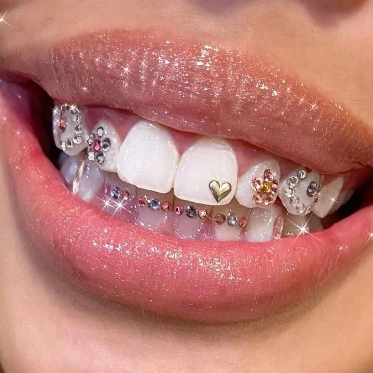 Nahaufnahme eines Lächelns mit weißem Gebiss, kunstvollen Zahnschmuck und glitzernden Effekten auf Lippen und Zähnen.
