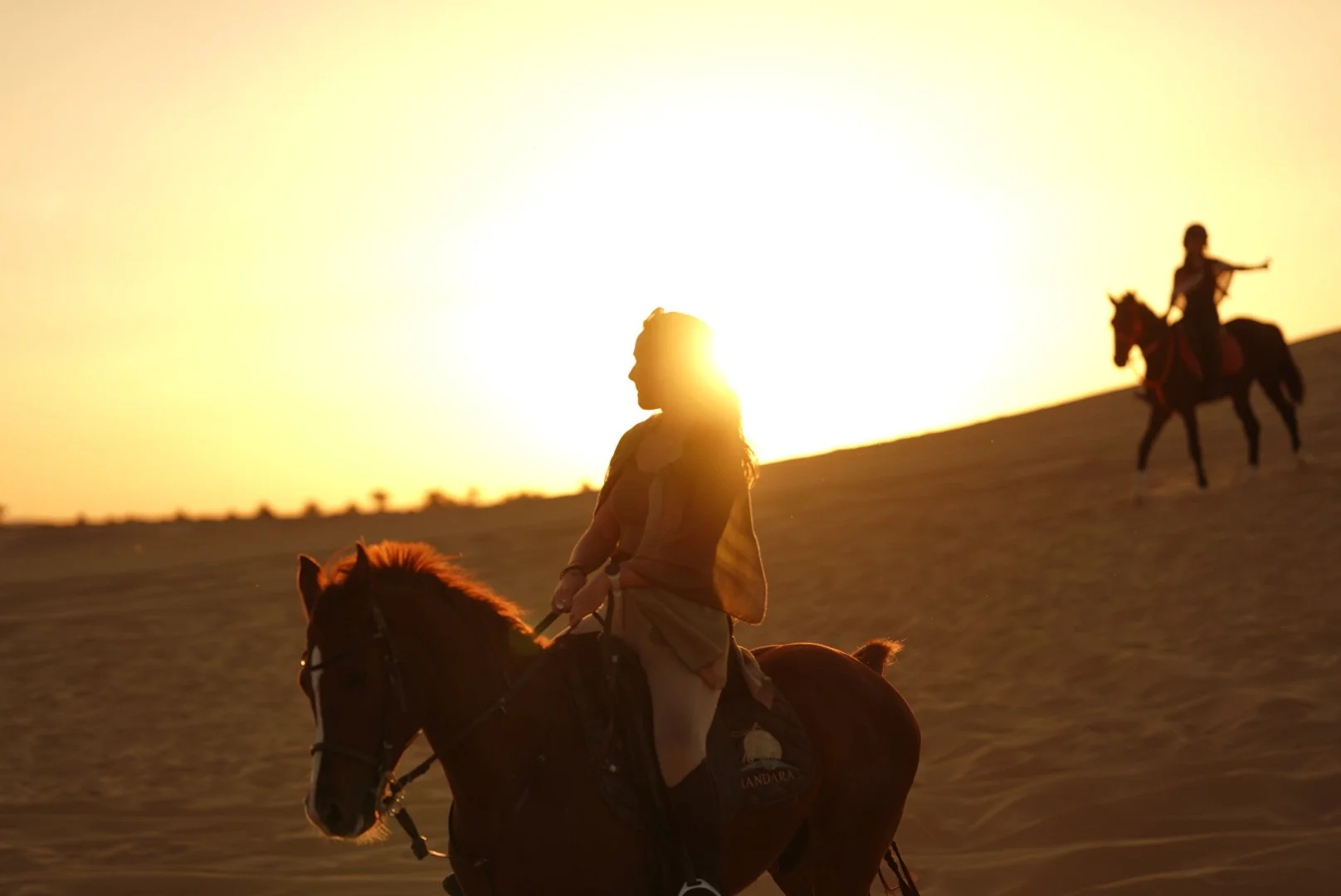 Silhouette of a woman riding a horse on a sand dune during sunset, with another rider on horseback in the background.