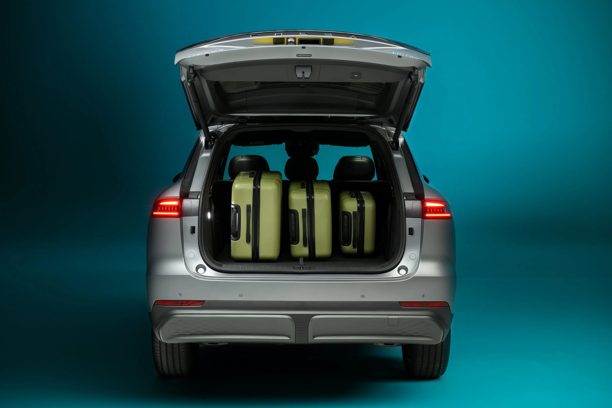 A silver SUV with its rear hatch open, revealing four light green suitcases inside on a solid teal background.