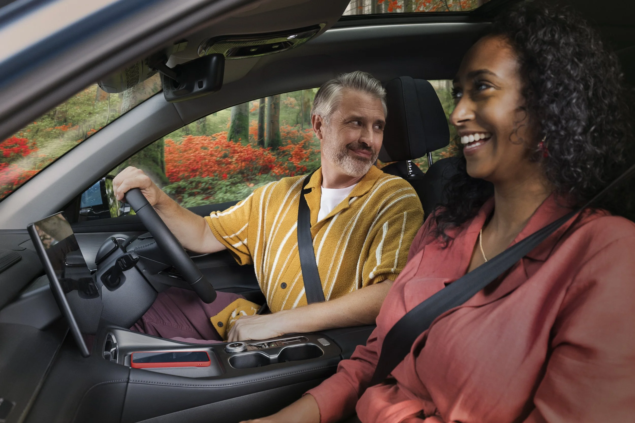 A man and woman sitting inside a car, smiling at each other. The man is in the driver's seat and the woman is in the front passenger seat. Outside the car, there are colorful autumn trees.